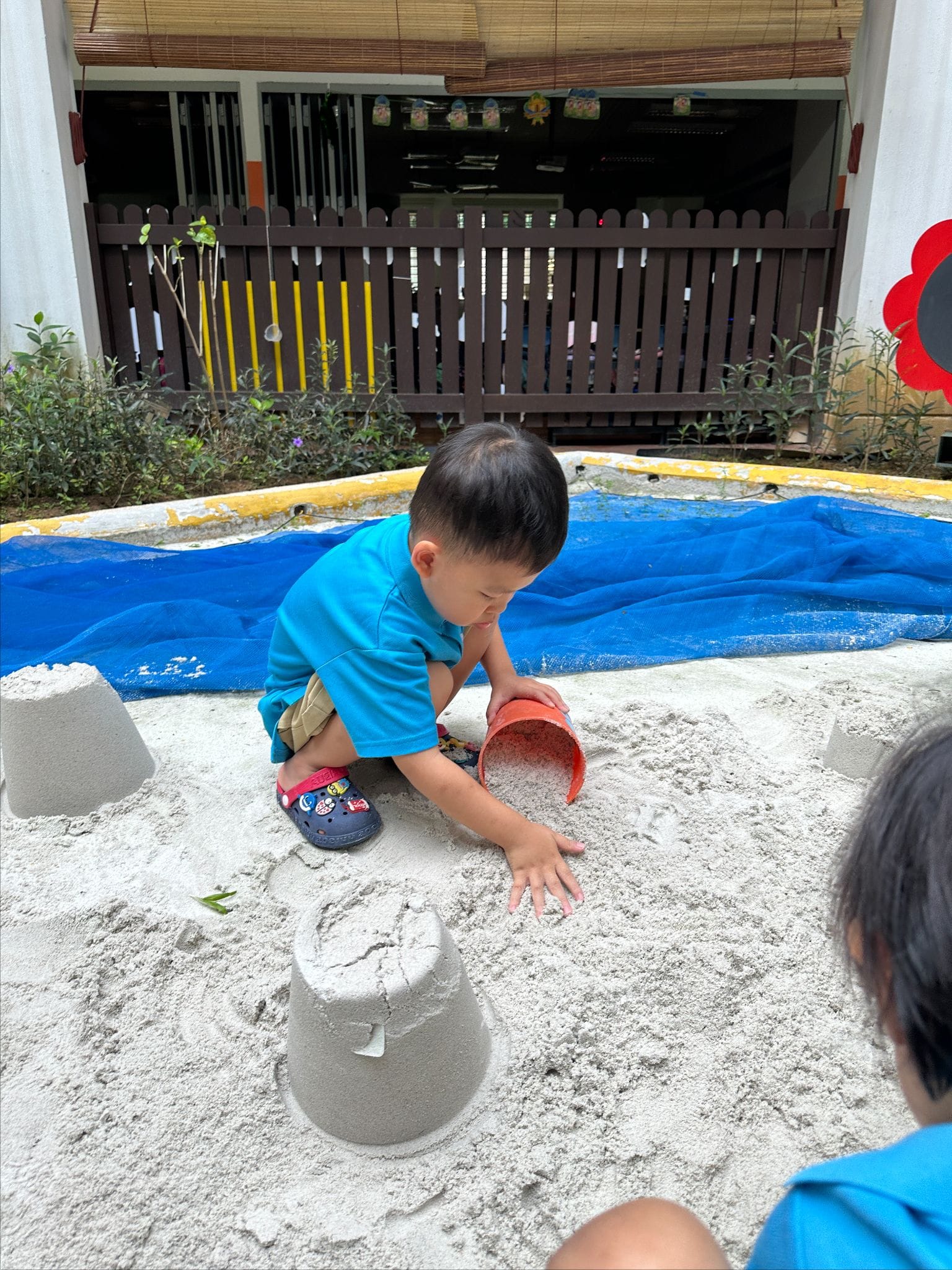 Young child in a blue shirt playing in a sandbox, tipping a red bucket near sand molds, outdoors.