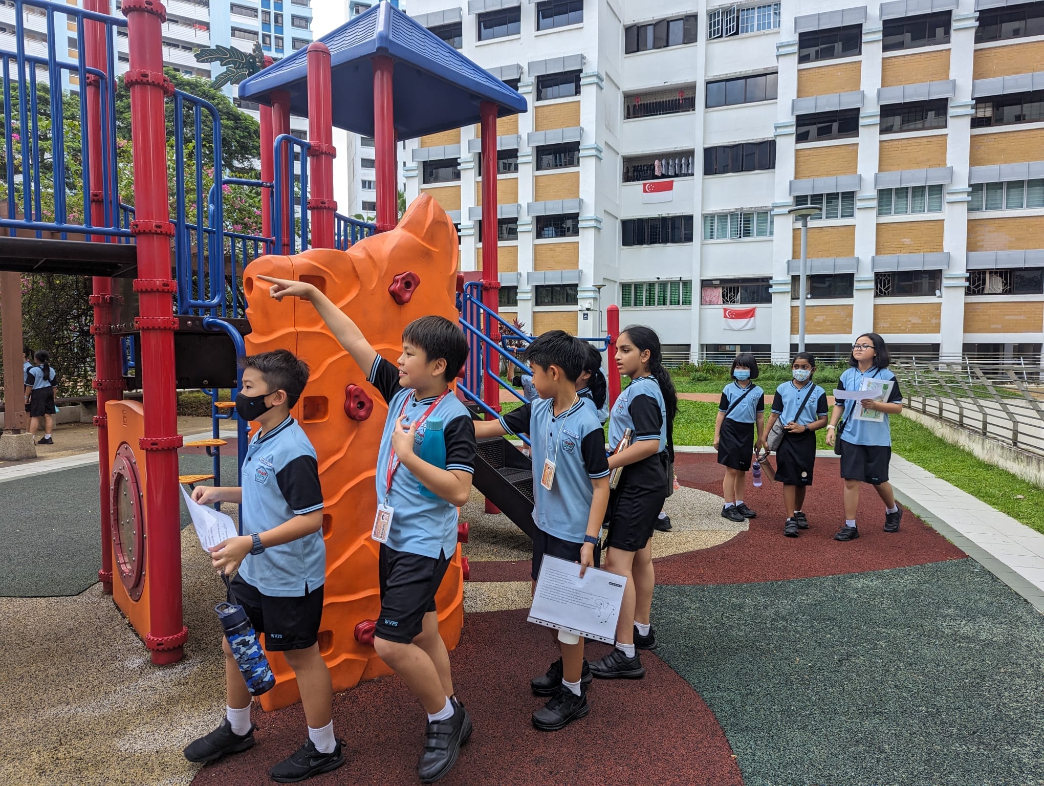 Several uniformed schoolchildren holding papers stand near a colorful orange and red playground structure beside a tall residential building.