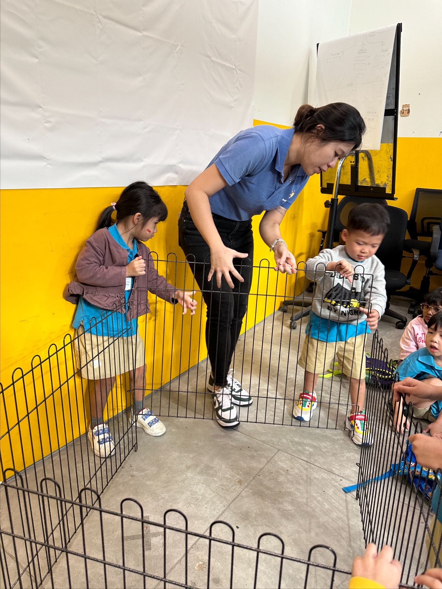 A woman in a blue shirt leans over a small metal pen, guiding two young children standing inside it.