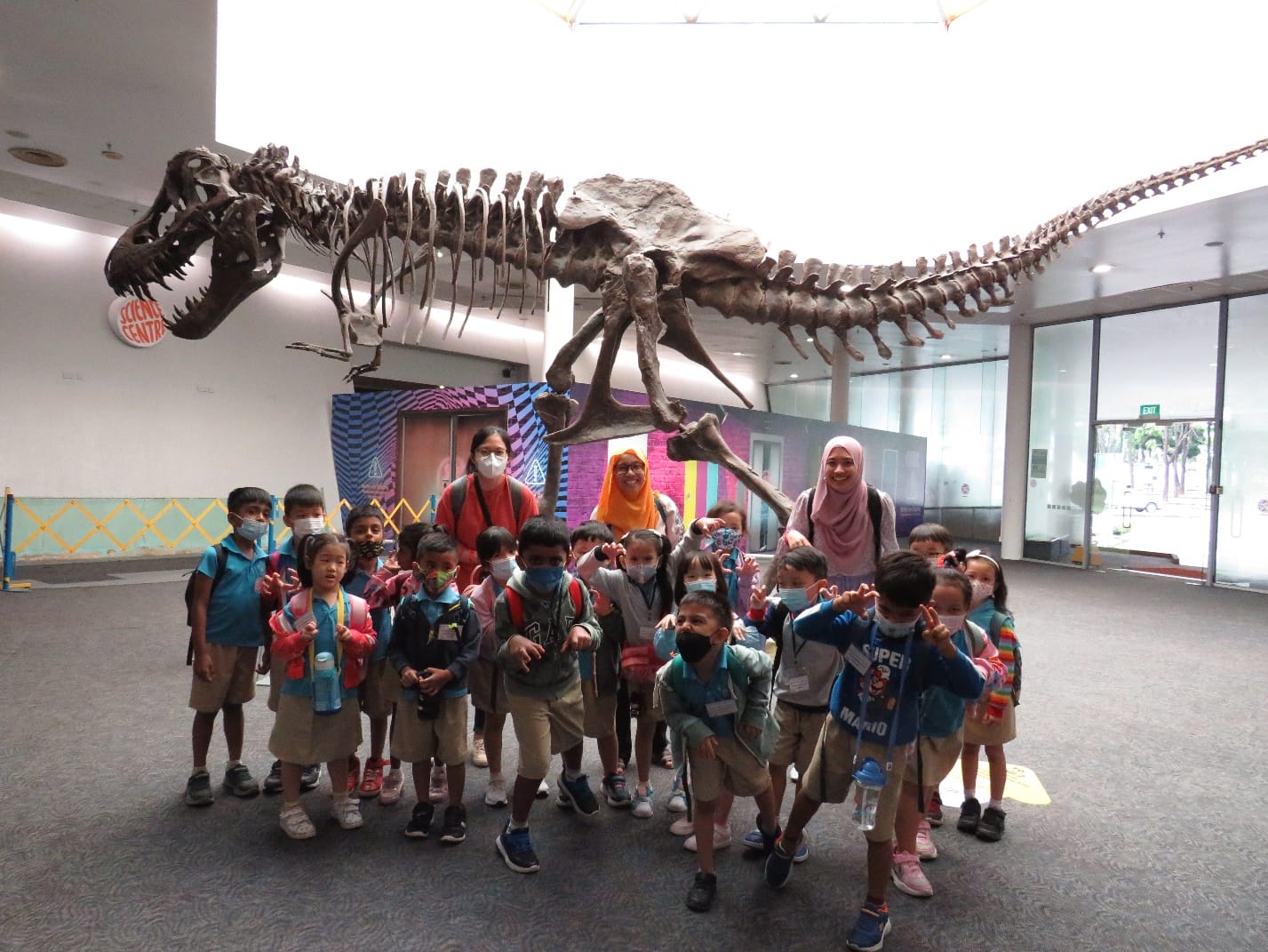 A group of young children and three adults pose in front of a large T-rex skeleton display at a Science Centre.