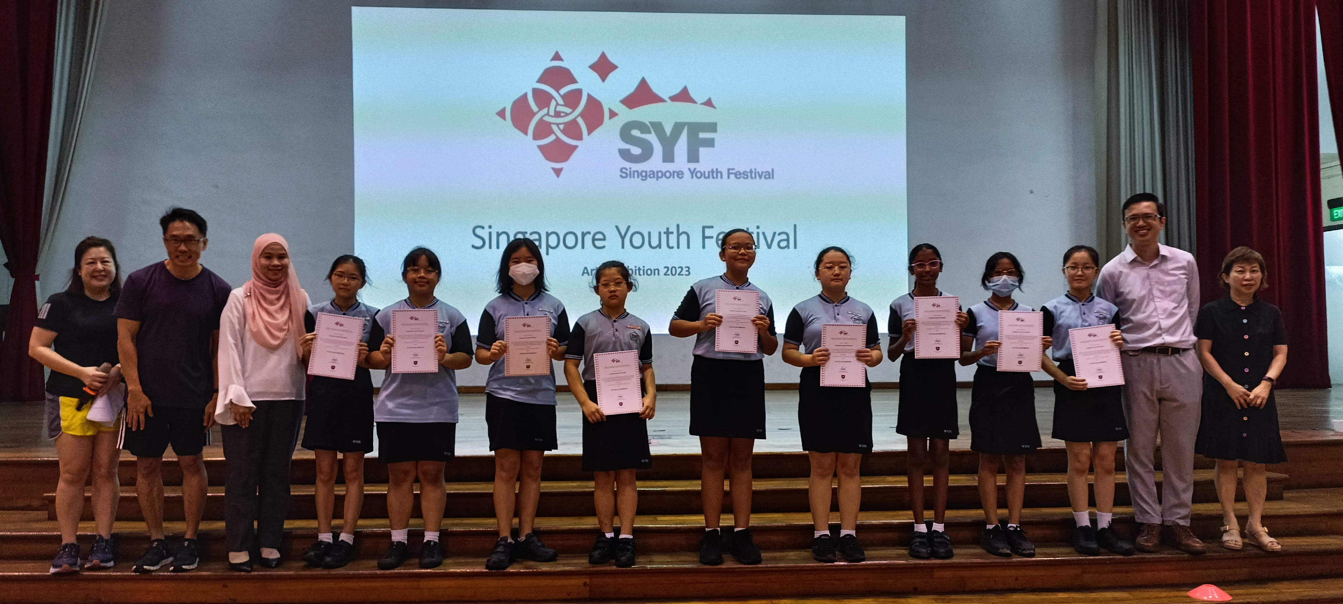 Students in uniforms hold certificates on a stage before a Singapore Youth Festival Art Exhibition 2023 screen.