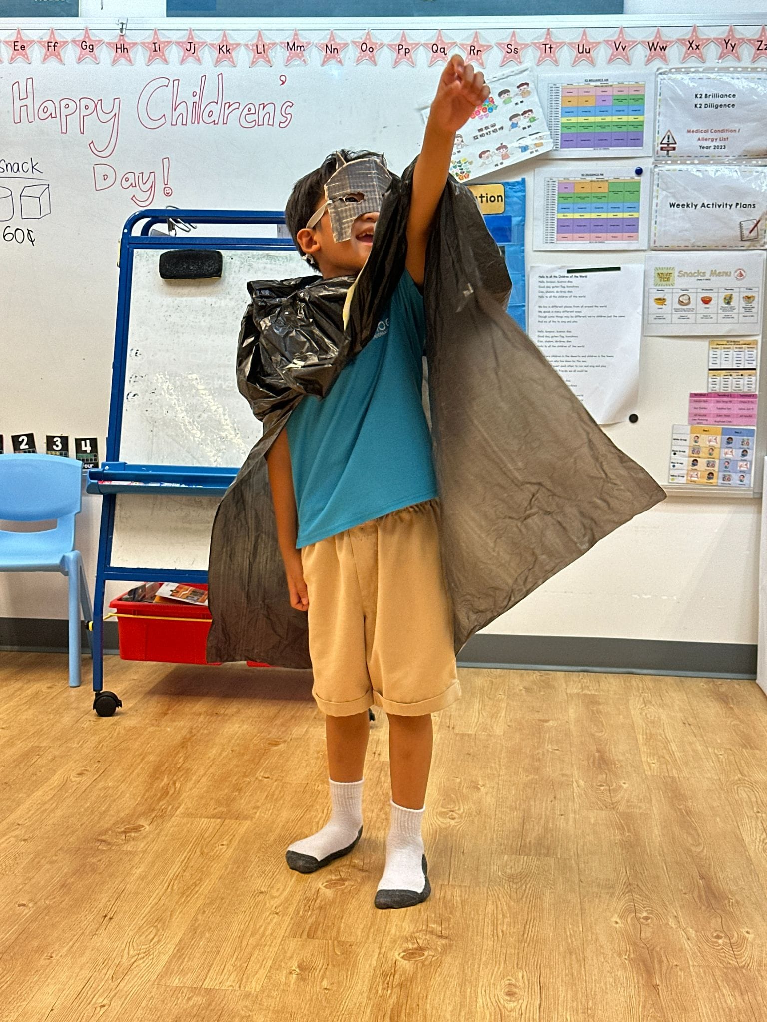 A young boy in a classroom wearing a homemade superhero costume with a black plastic bag cape and mask, fist raised.