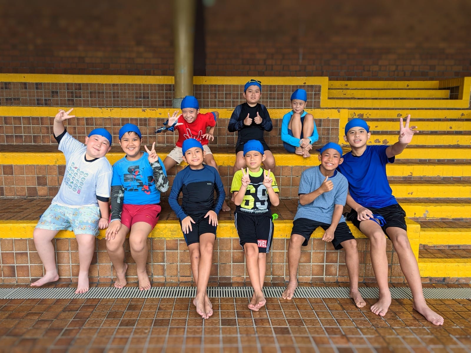 Nine boys wearing blue swim caps sit on yellow-trimmed tiled bleachers, smiling and making peace signs.