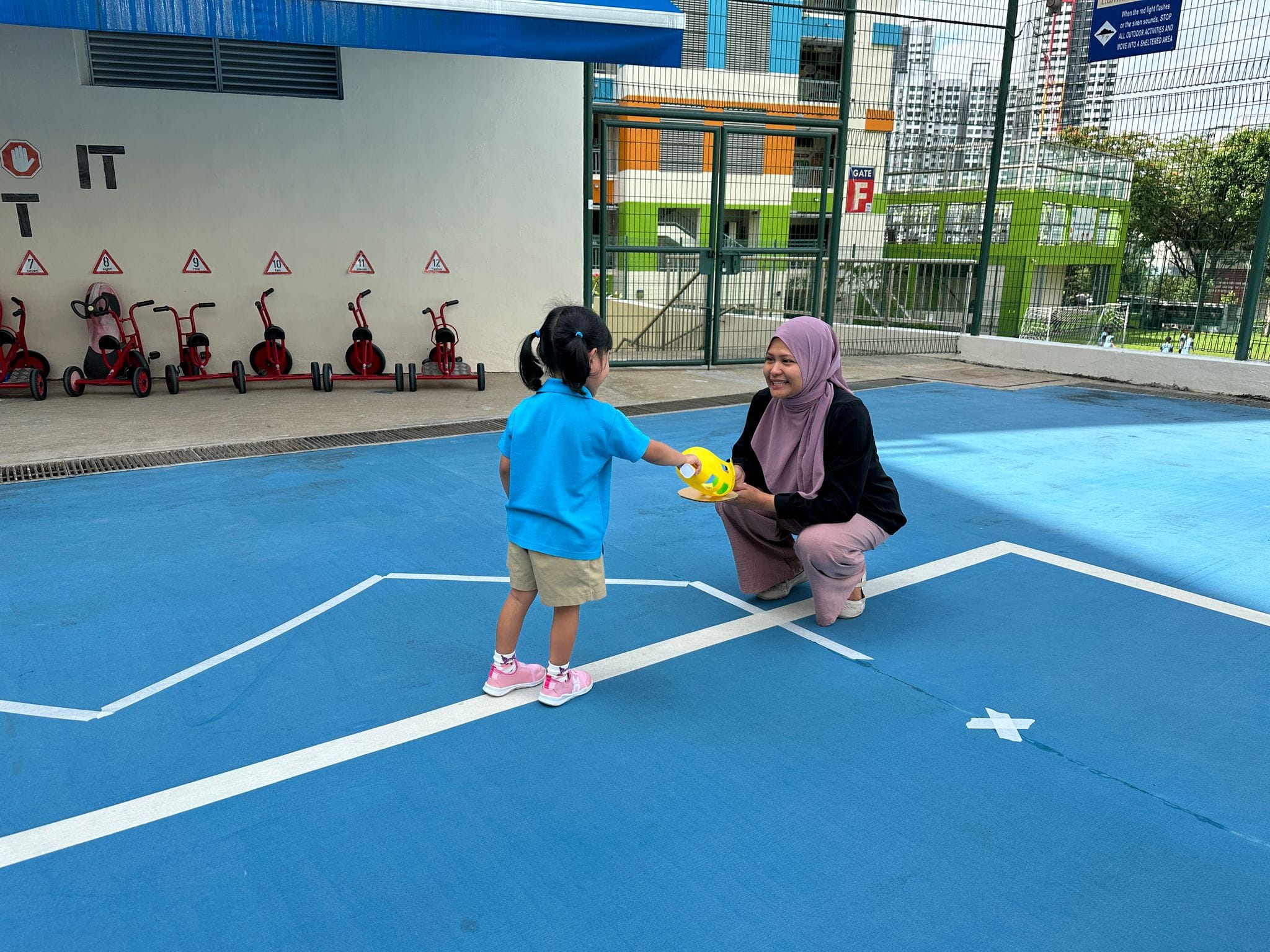 A woman in a hijab crouches smiling as a young girl in a blue shirt hands her a yellow toy on a school playground.
