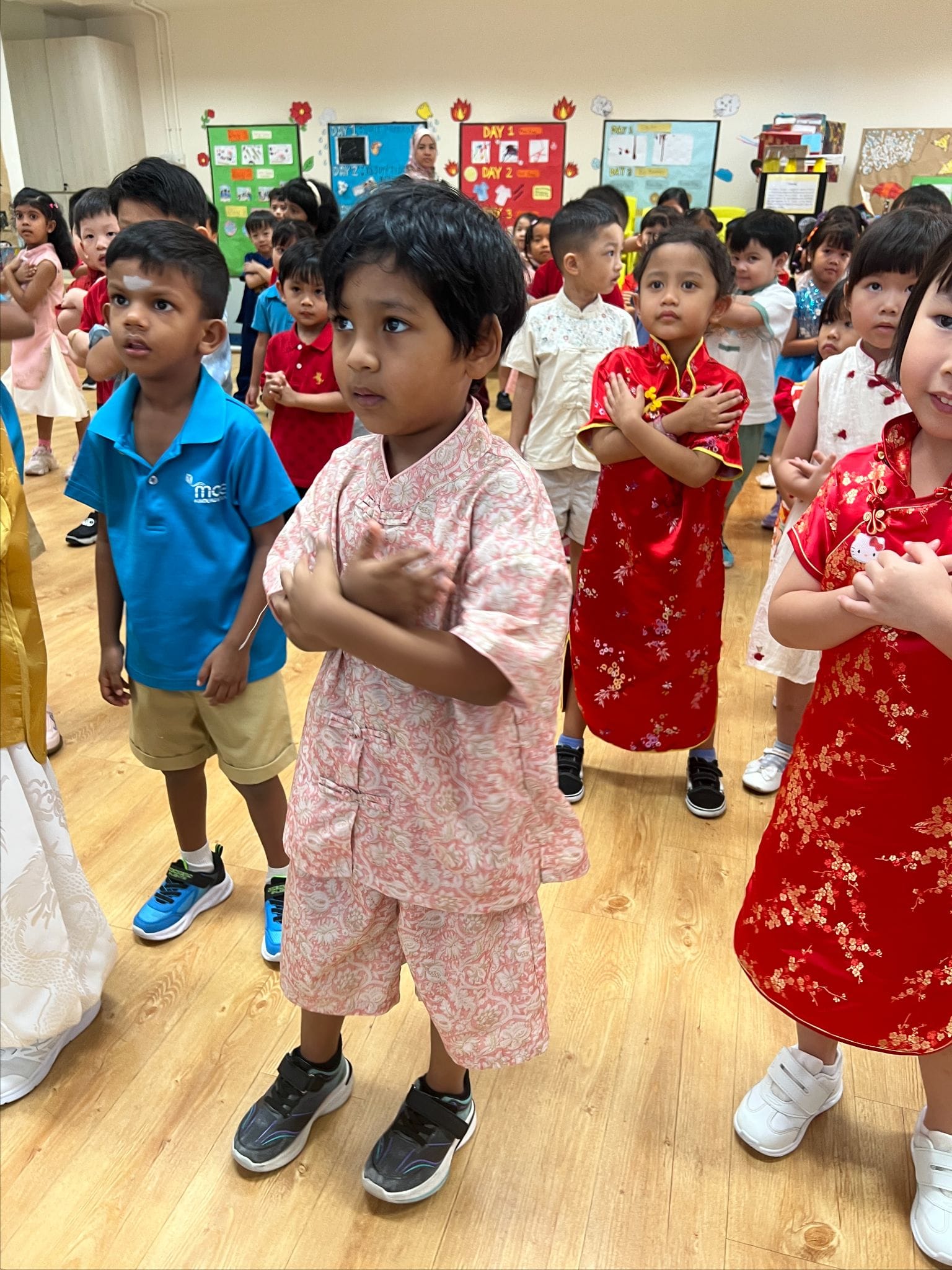 Young children in traditional outfits, including red cheongsams and batik, stand in a decorated classroom.