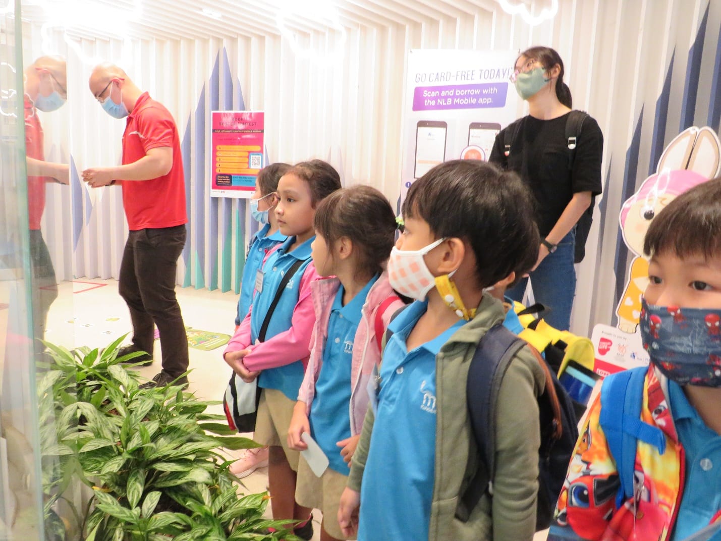 Children in blue uniforms and face masks line up indoors near an NLB mobile app sign, with two adults nearby.