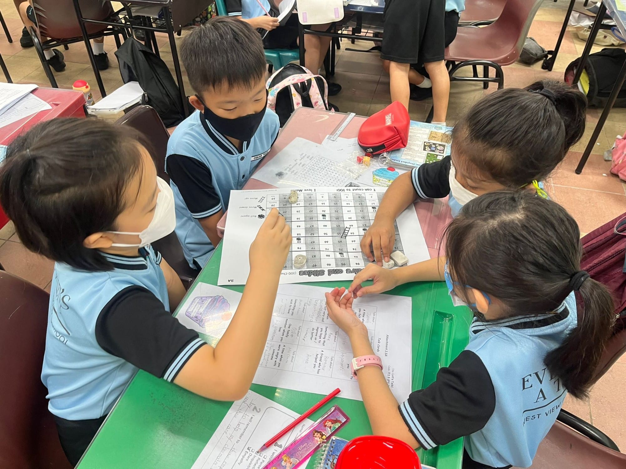 Four young students in blue uniforms work together on a Snakes and Ladders math worksheet at a green classroom table.