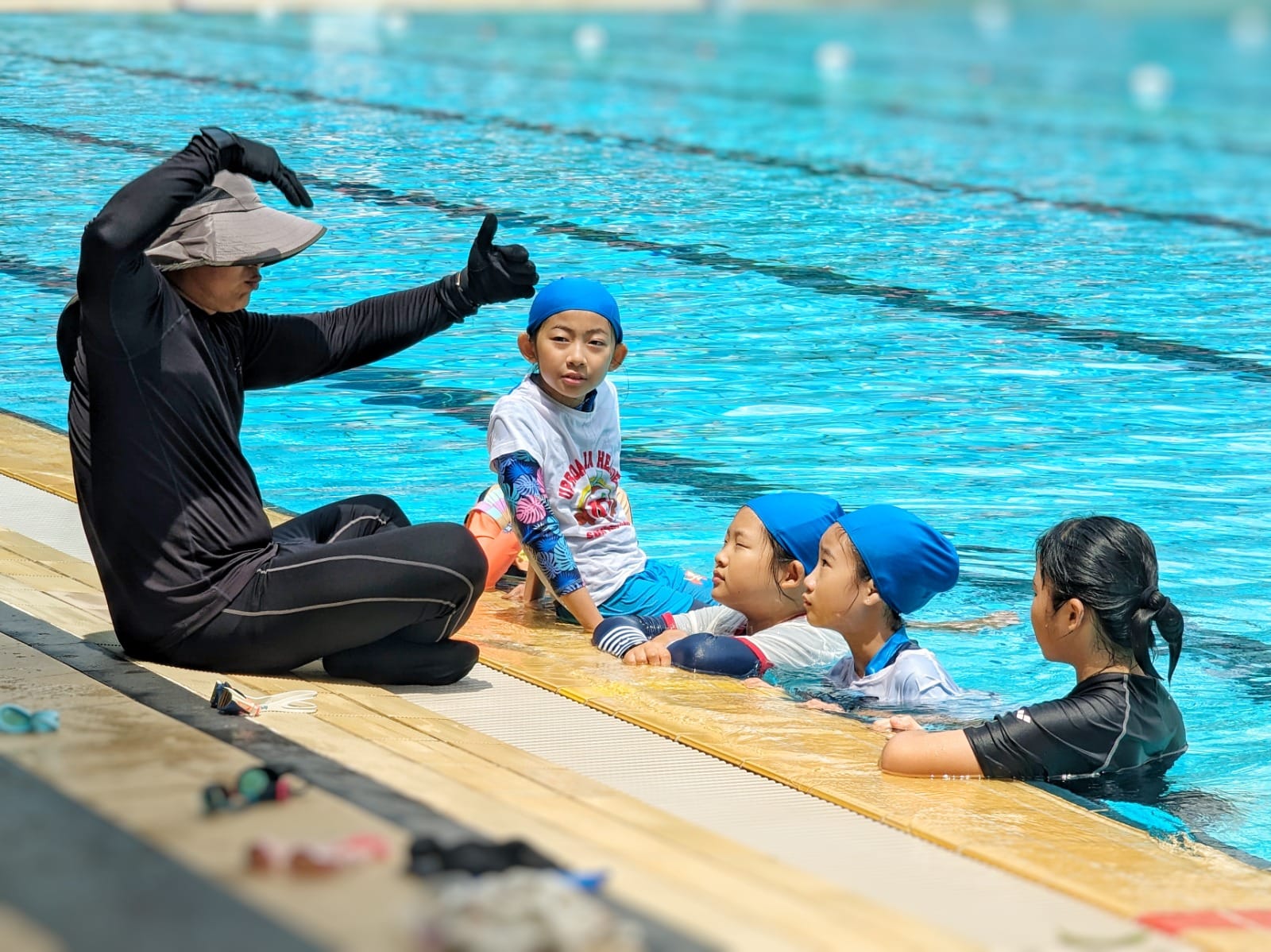 A swim instructor in black sits poolside gesturing to three young children in blue caps at the pool edge.