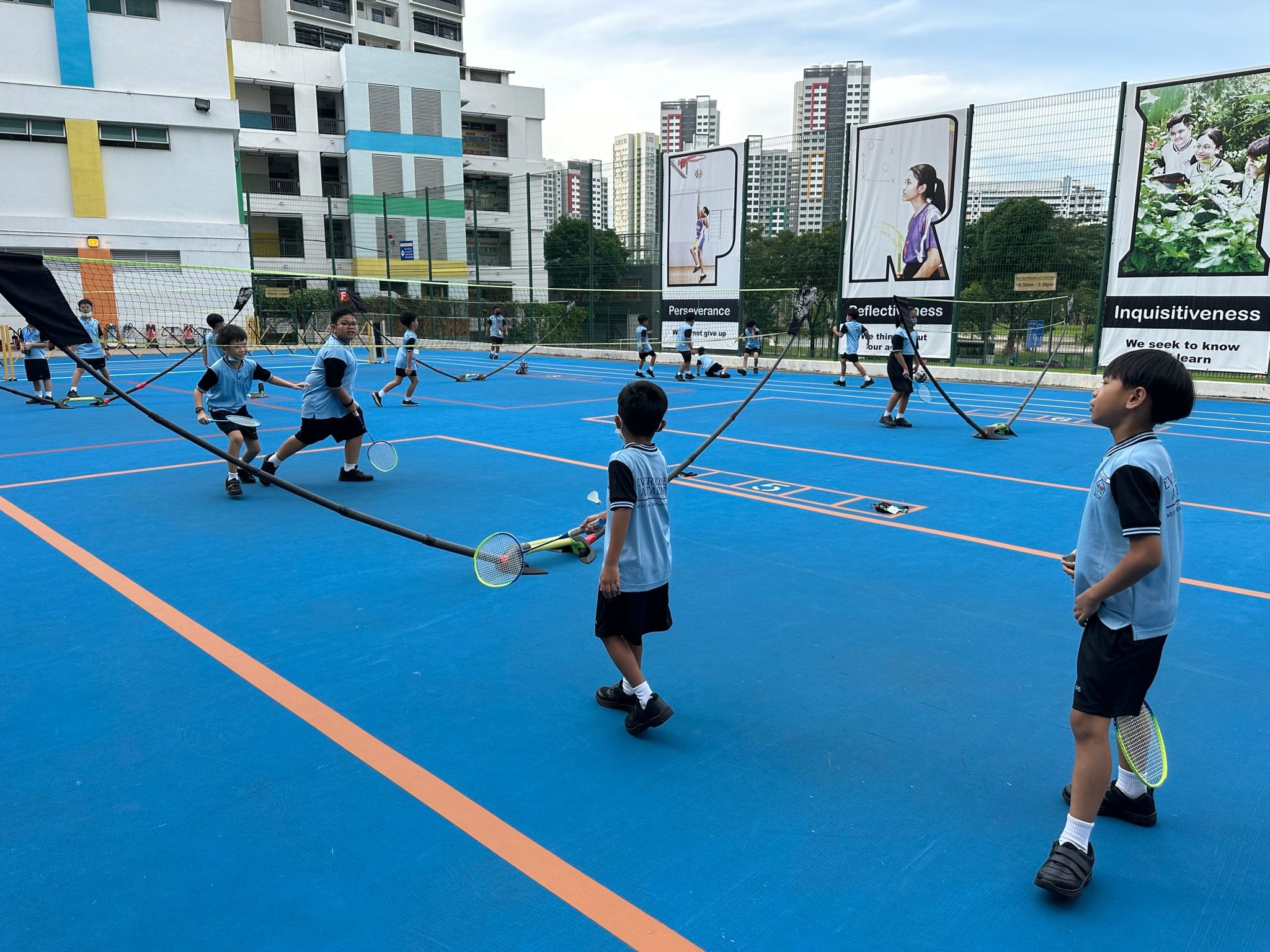 Children in school uniforms play badminton on an outdoor blue court with nets, surrounded by school banners and buildings.