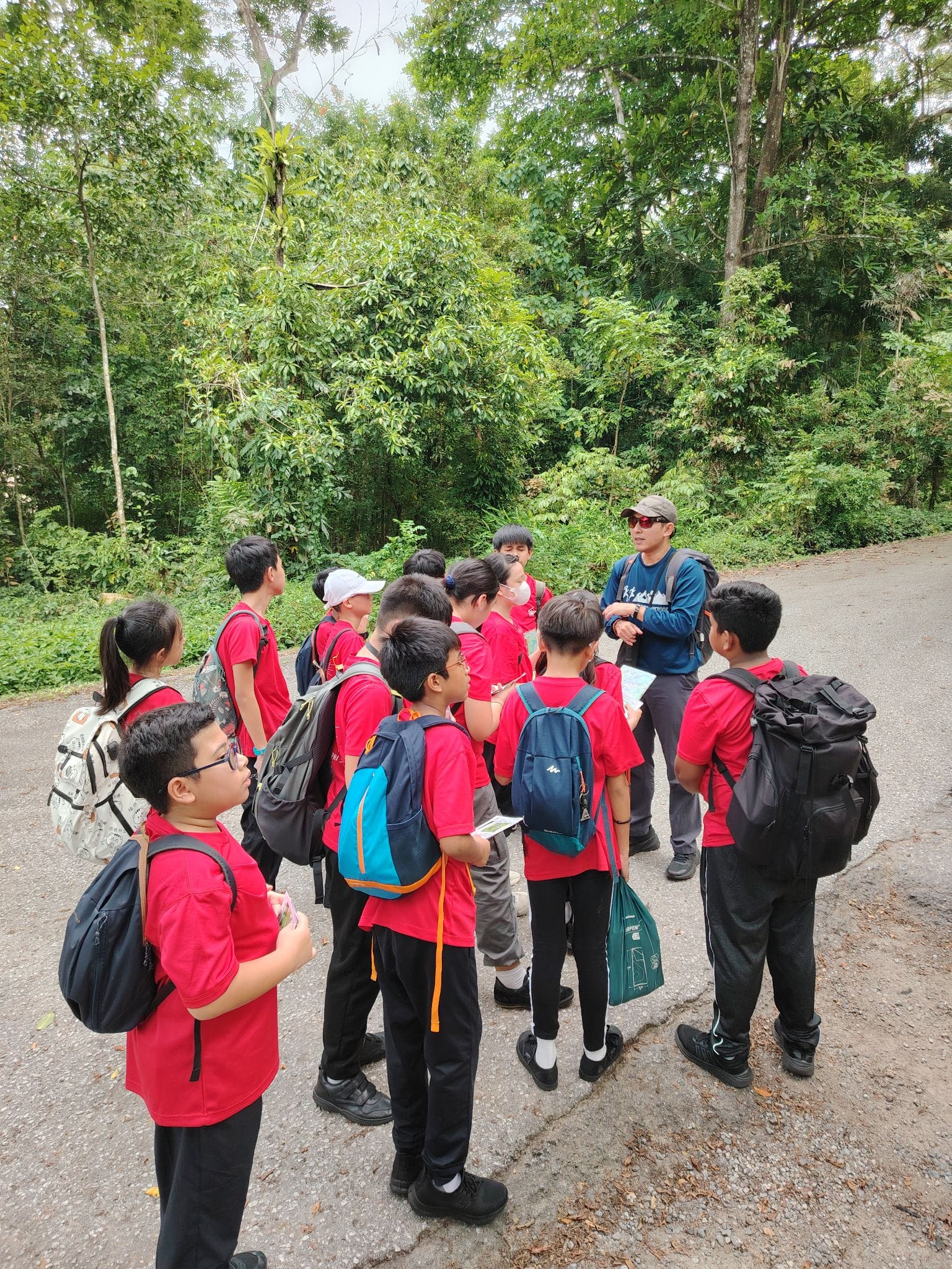 A group of children in red shirts with backpacks listen to an adult guide on a forest path.