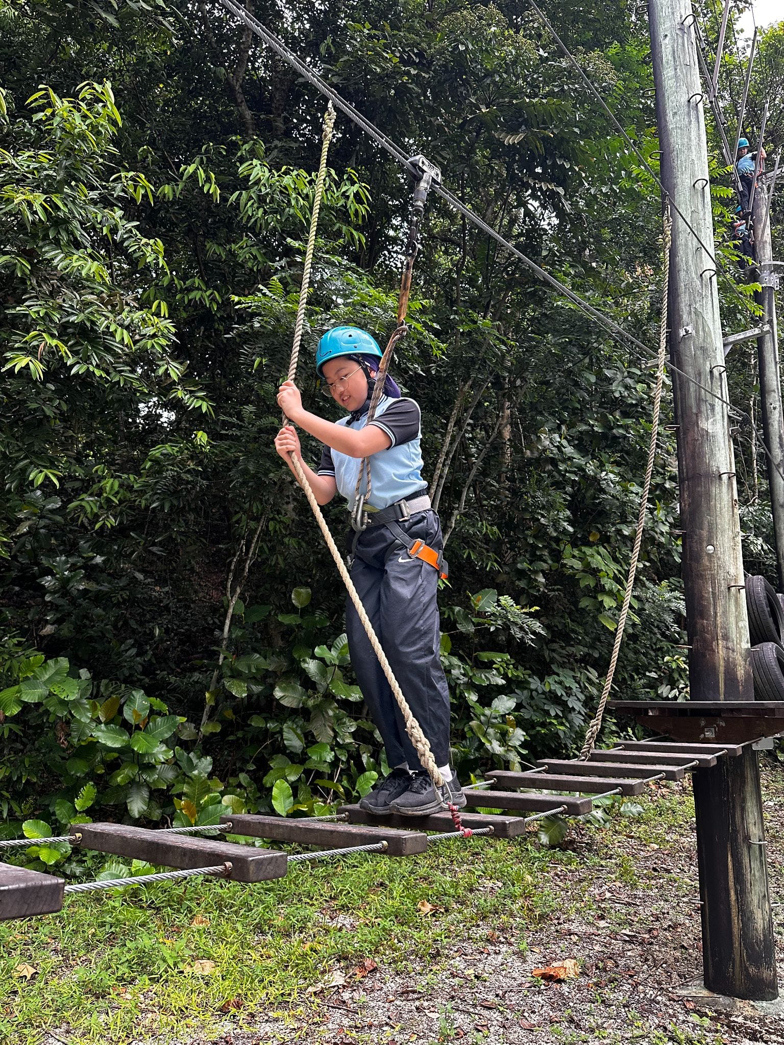 A child wearing a blue helmet and safety harness walks across a suspended wooden rope bridge outdoors.