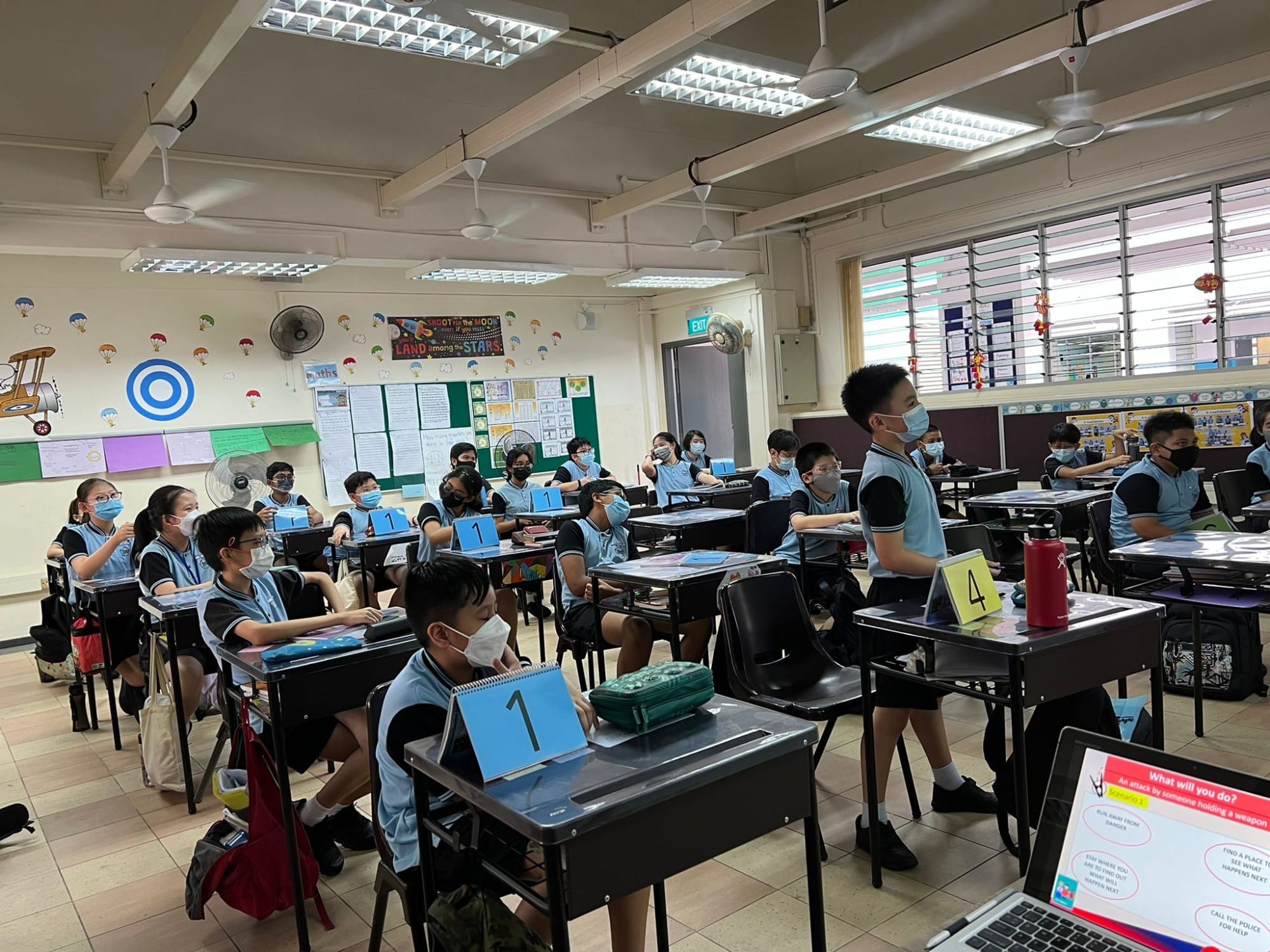 Students in blue uniforms and face masks sit at numbered desks in a decorated classroom during a lesson.
