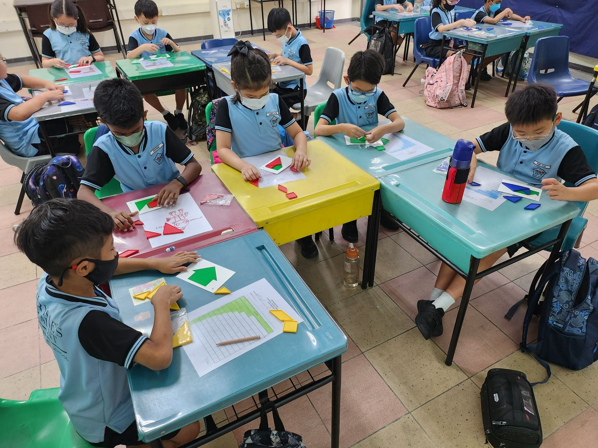 Young students in blue uniforms and masks sit at colorful desks arranging geometric shape pieces on worksheets.