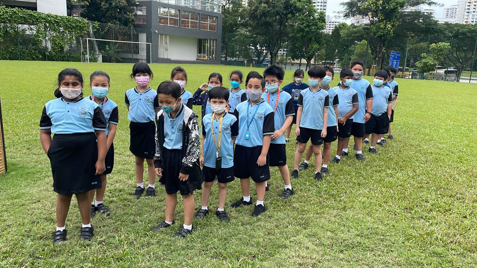 A group of young students in blue and black uniforms wearing face masks stand on a grassy school field.