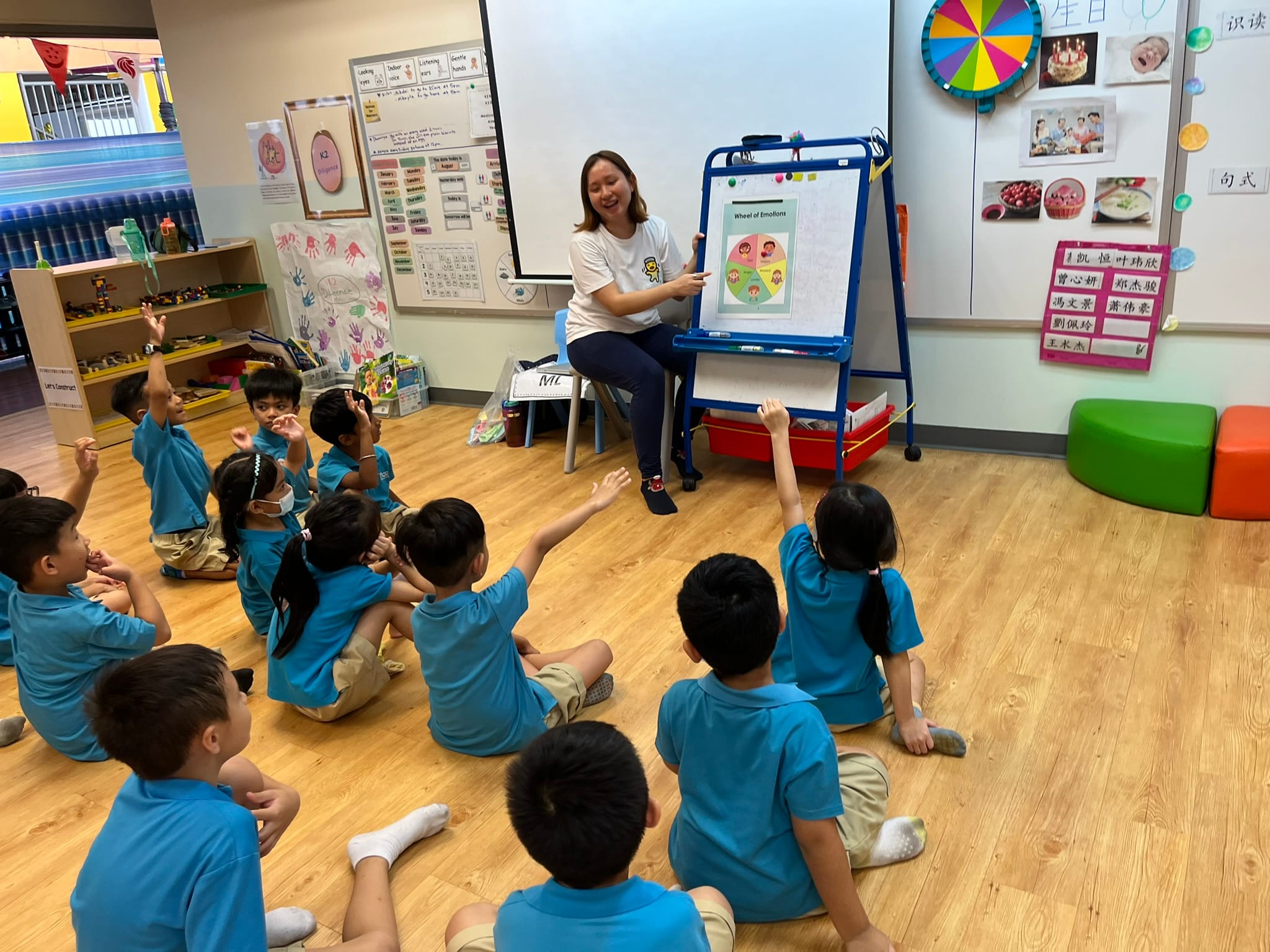 Teacher sits beside an easel showing a Wheel of Emotions chart while young children in blue uniforms sit on the floor raising hands.