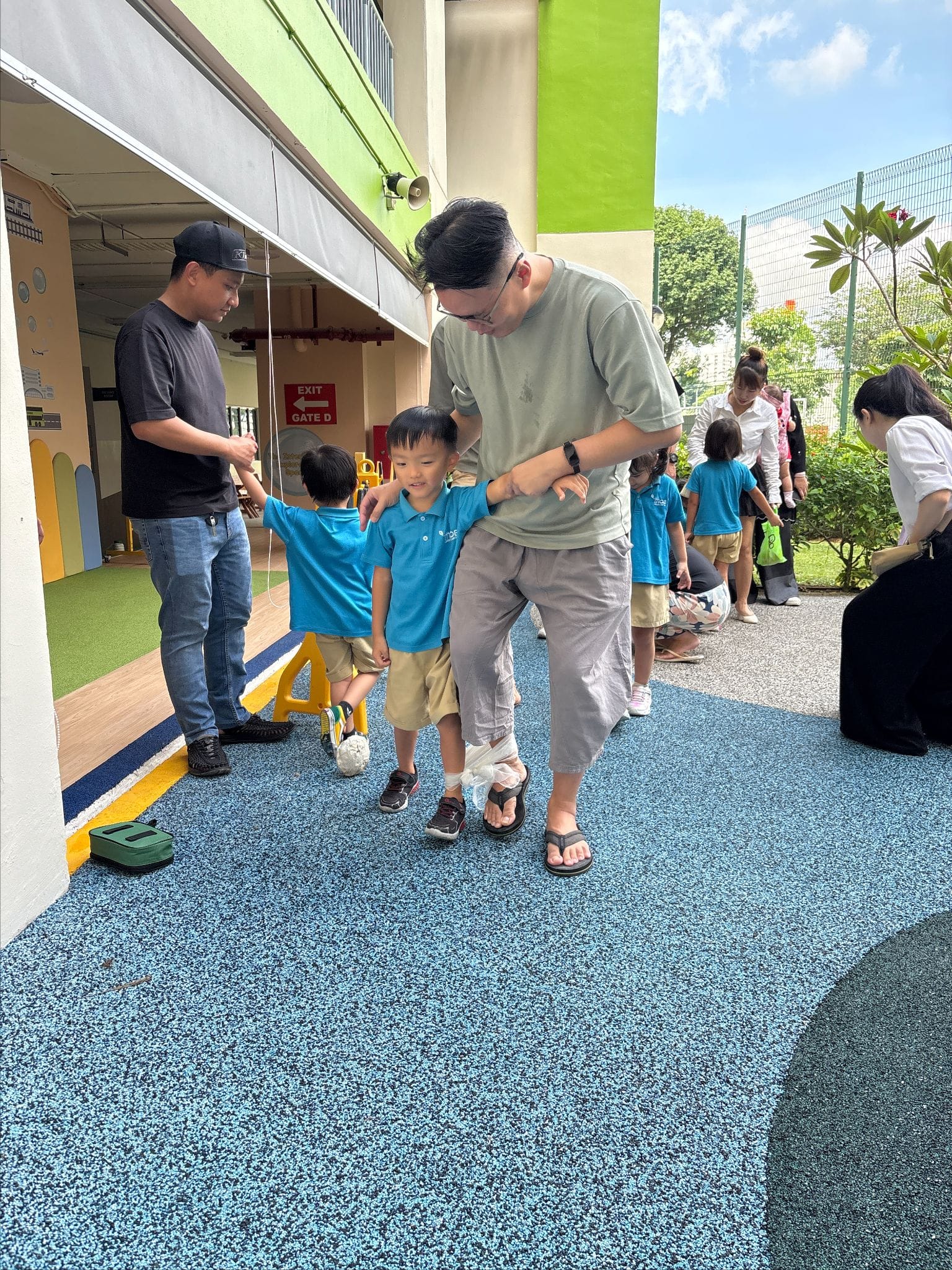 An adult bends down to guide a young child in a blue shirt outside a colorful school building.