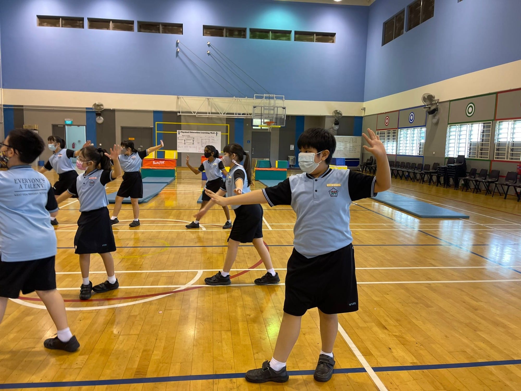 Children in blue school uniforms and face masks doing exercises in a school gymnasium with wooden floors.