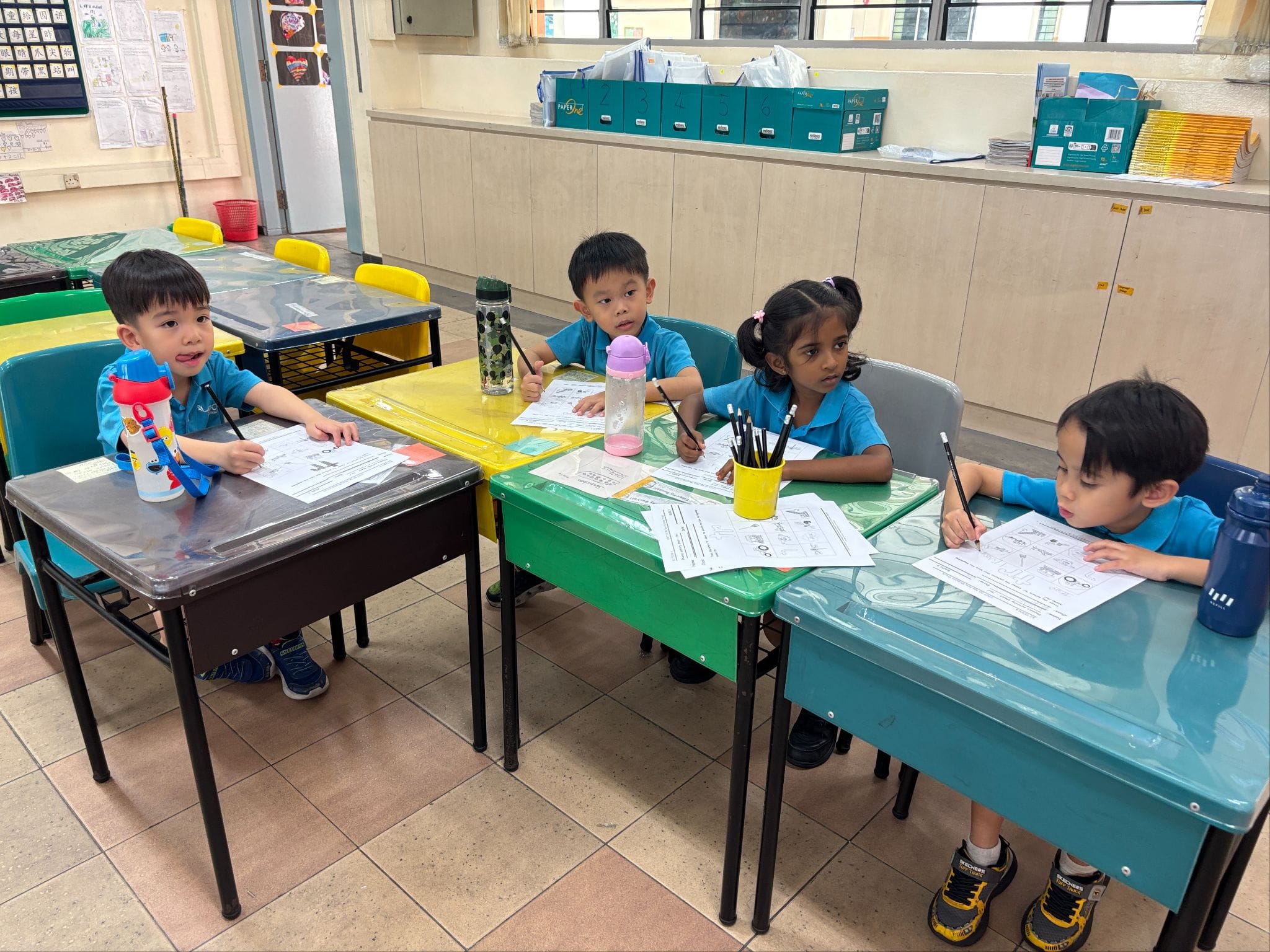 Four young children in blue uniforms sit at colorful desks in a classroom, working on worksheets with pencils.