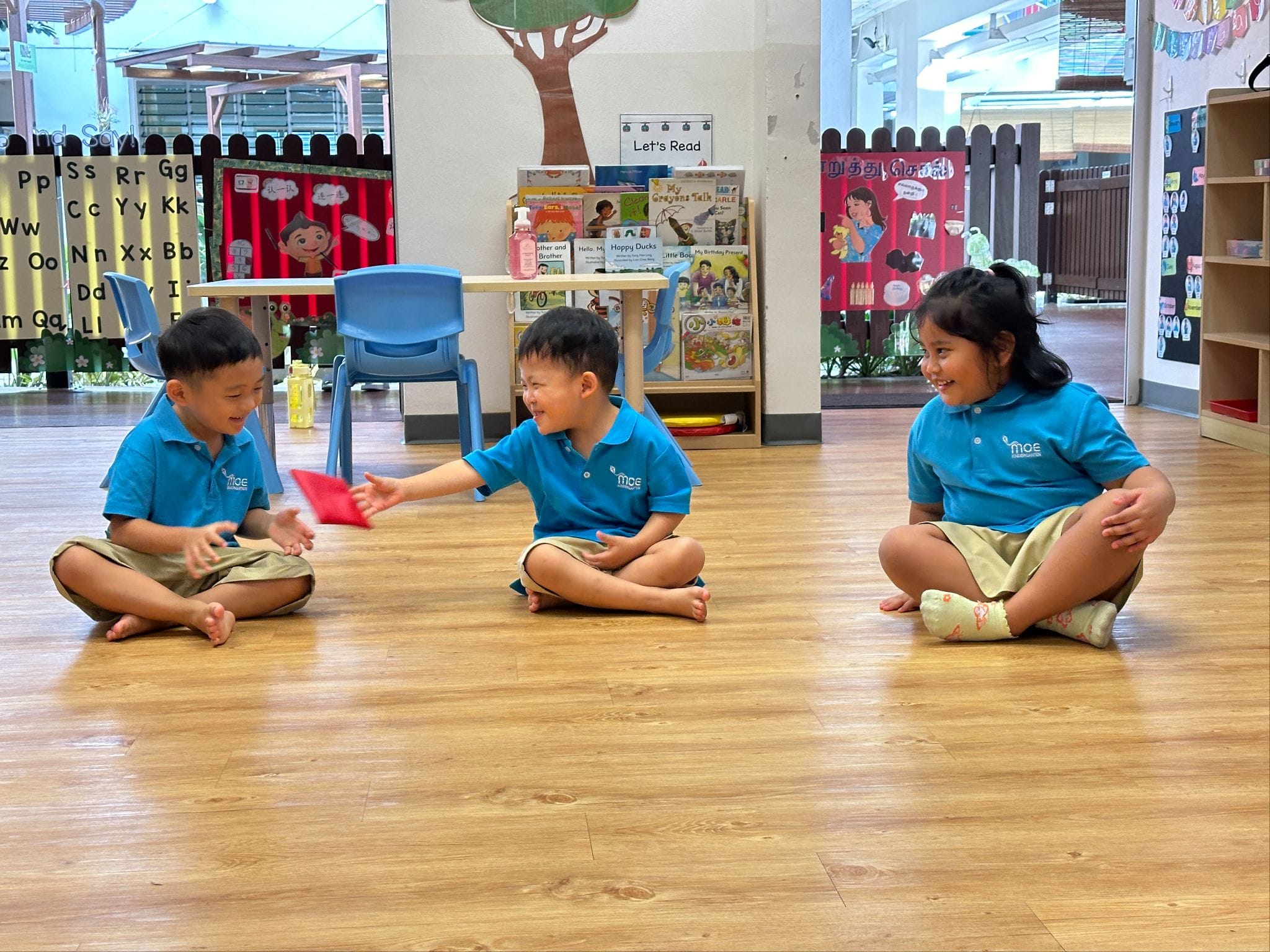 Three young children in blue uniforms sit cross-legged on a classroom floor, tossing a red beanbag to each other.