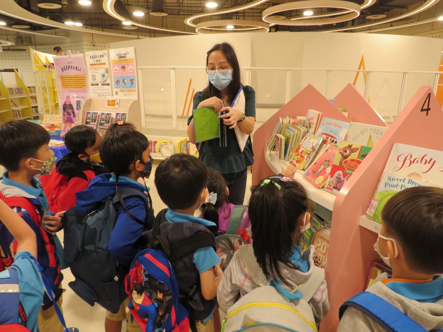 A masked woman holds a book while young children with backpacks gather around a colorful book display in a library.