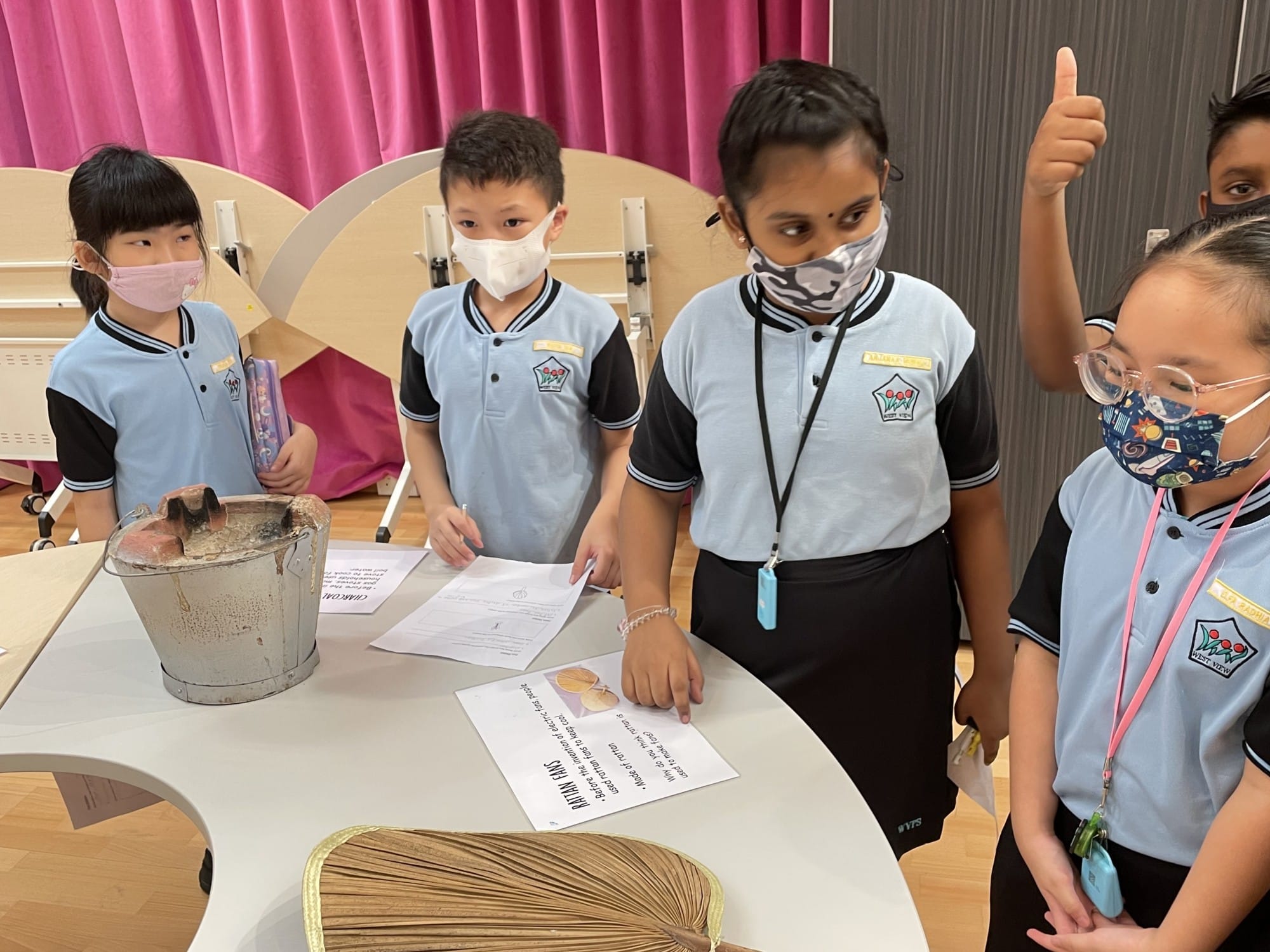 Four masked school children in blue uniforms stand around a table with a bucket, papers, and a fan, one giving thumbs up.