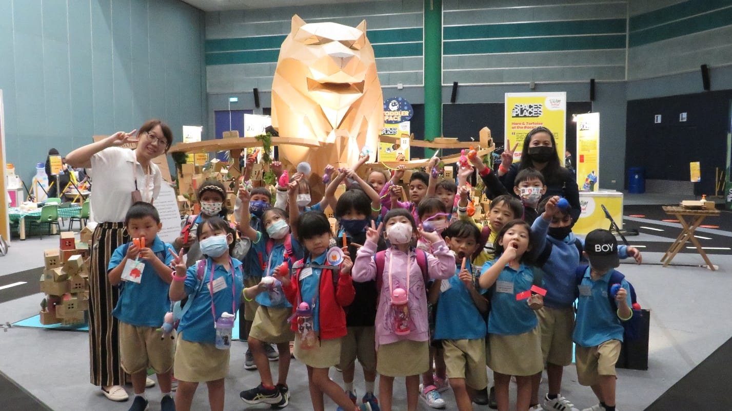 A group of young children in school uniforms pose with two adults before a large geometric lion sculpture indoors.