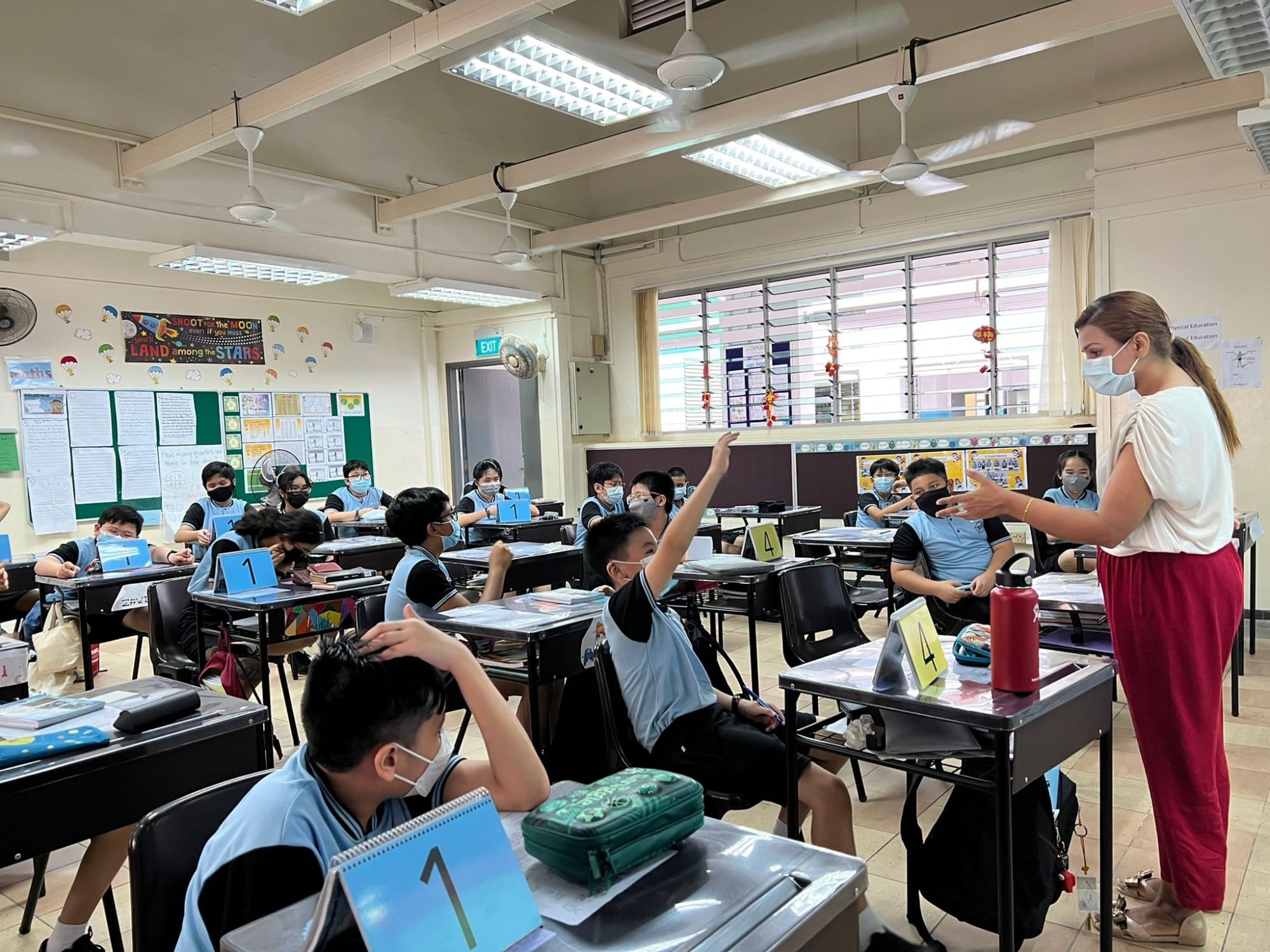A masked teacher stands at the front of a classroom while students in blue uniforms sit at desks, one raising a hand.