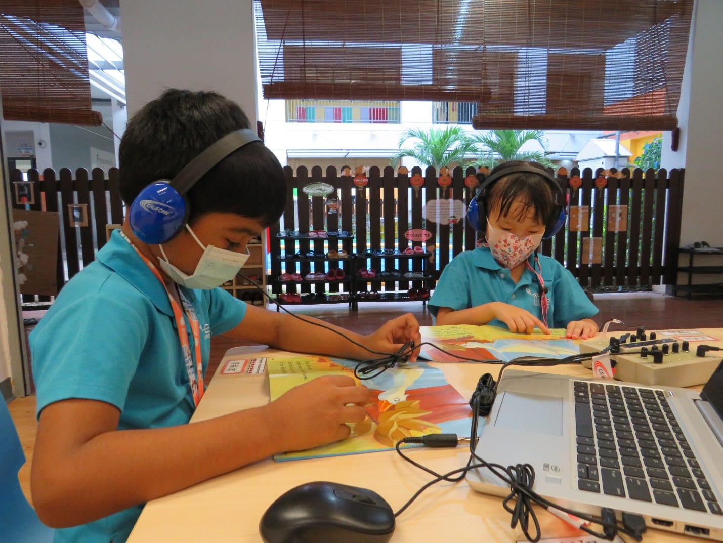 Two masked children wearing headphones read colorful books at a table with a laptop and audio equipment.