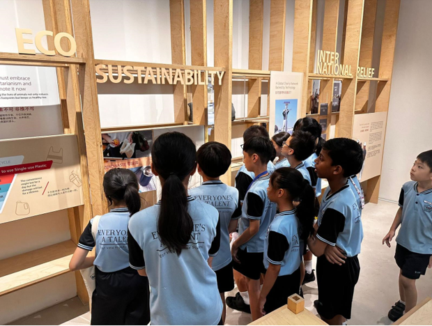 Children in light blue uniforms view a wooden exhibit display with signs reading Sustainability, Eco, and International Relief.