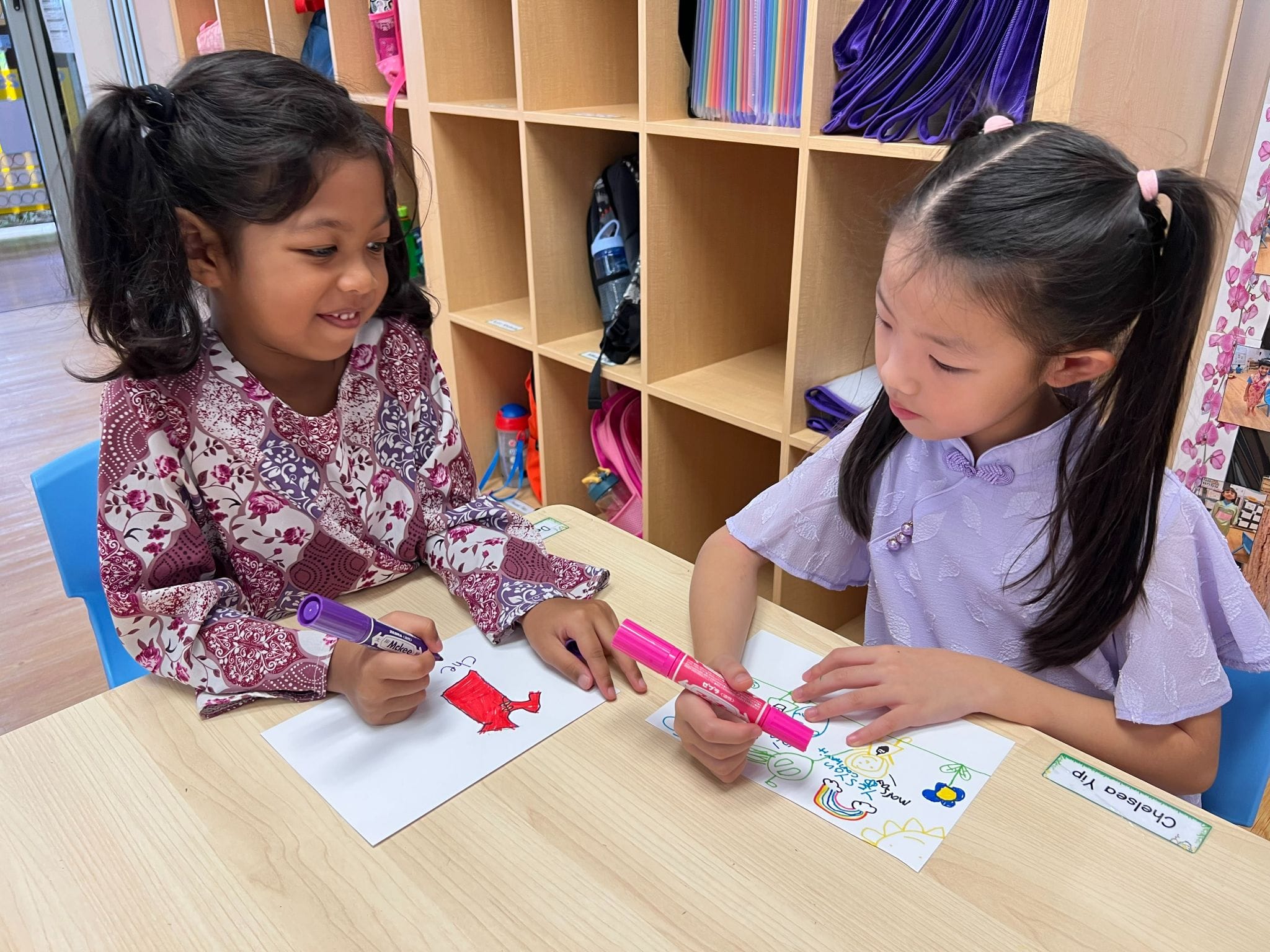 Two young girls sit at a classroom table drawing with colorful markers on white paper.