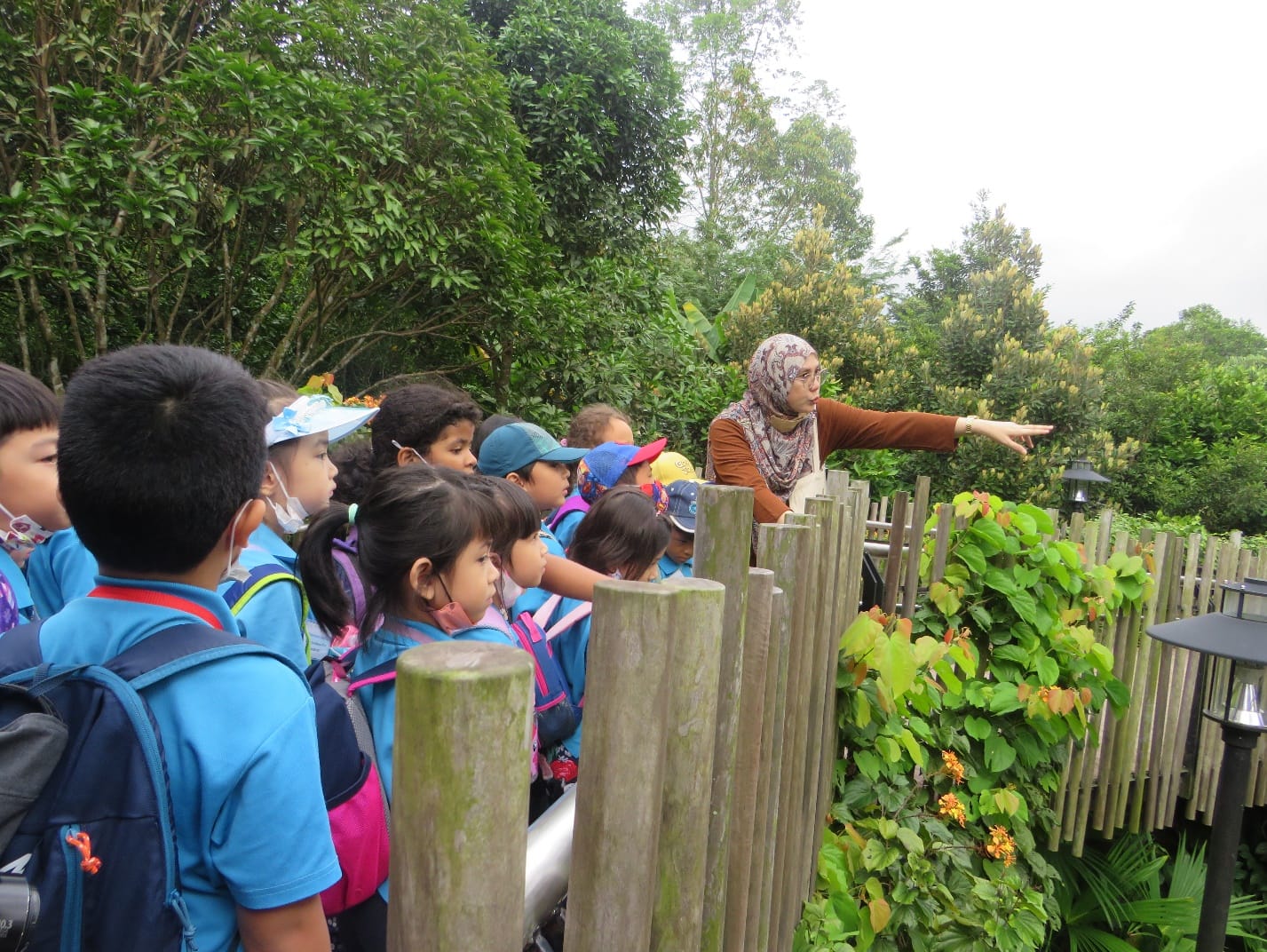 A woman in a hijab points at something beyond a wooden fence while young children in blue uniforms look on outdoors.
