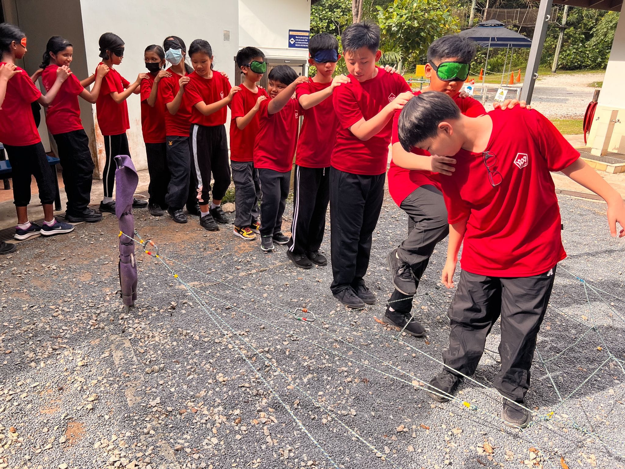 Children in red shirts form a line, some blindfolded, navigating a rope obstacle course on gravel outdoors.