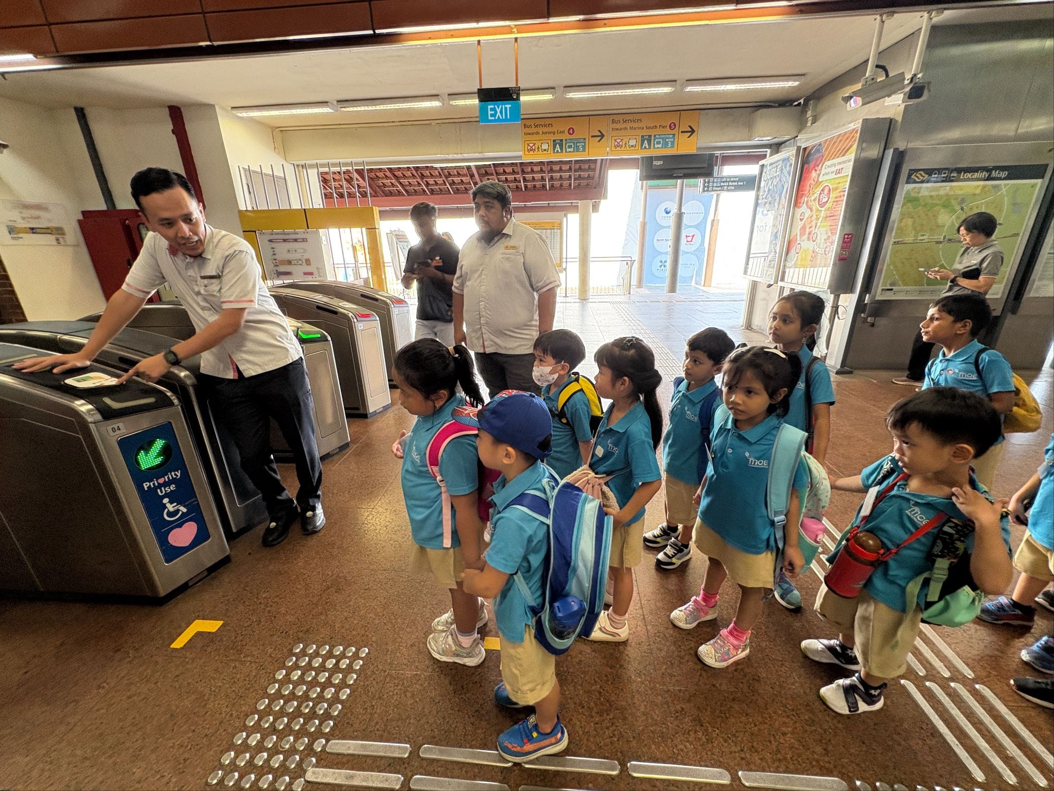 A group of young students in blue uniforms line up at MRT fare gates assisted by a station staff member.