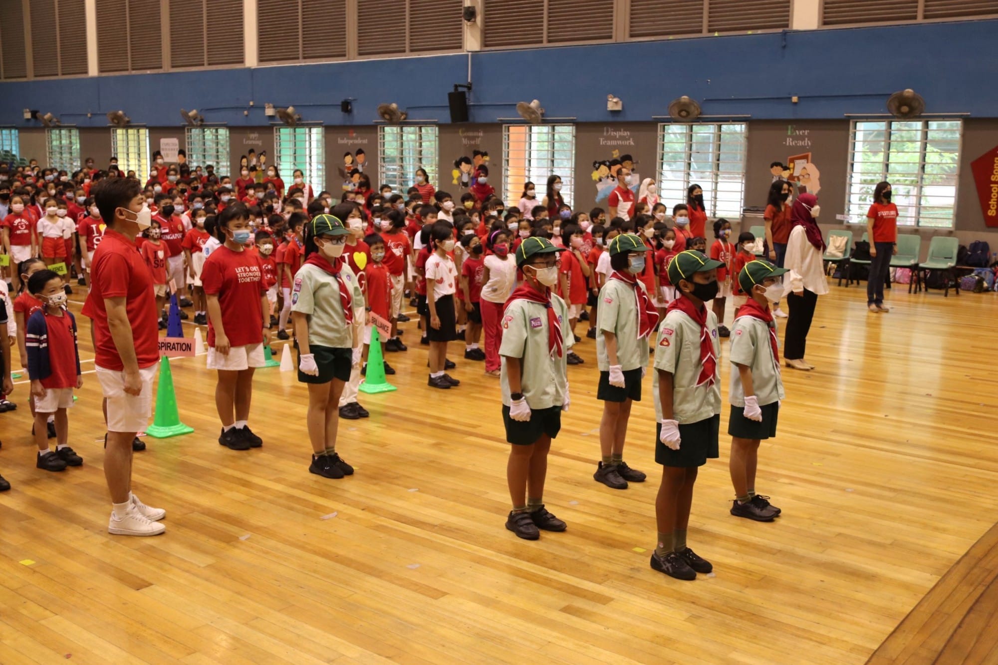 Children in scout uniforms and red shirts stand in rows in a school hall during an assembly.