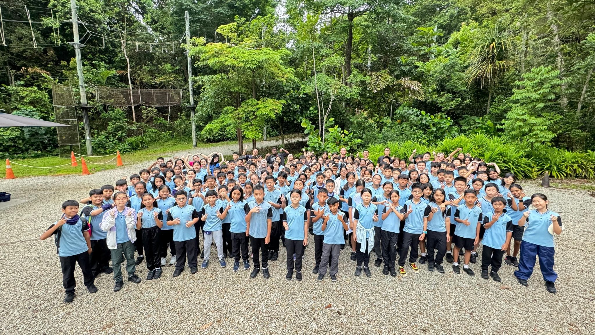 Large group of children in matching light blue uniforms posing outdoors on gravel, surrounded by dense green trees.