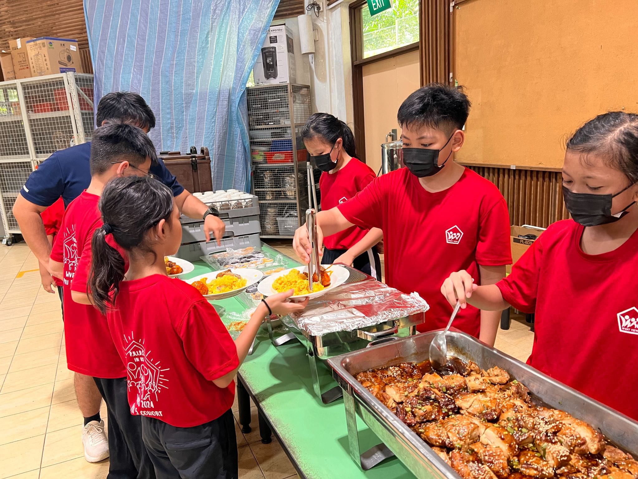 Students in red shirts and face masks serve food including rice and glazed chicken at an indoor buffet table.