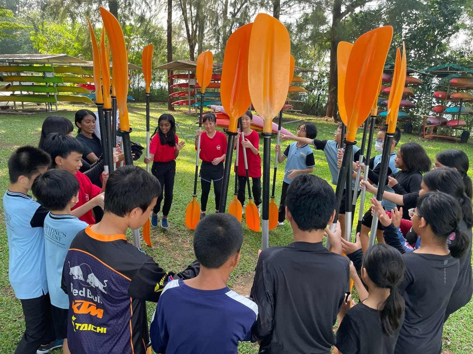 A group of children stand in a circle outdoors, each holding orange kayak paddles upright, with kayak racks visible behind them.