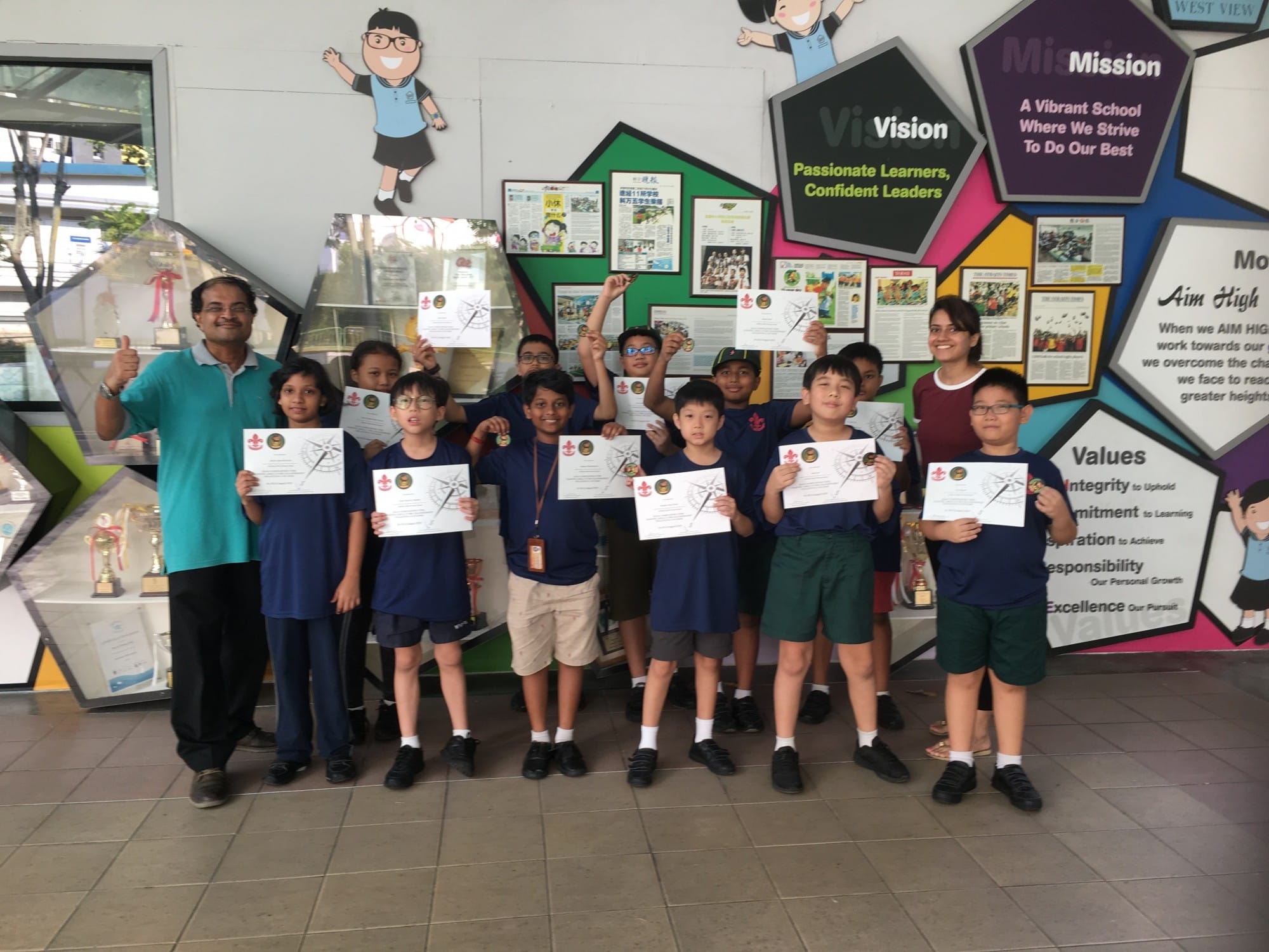 A group of young students holding certificates pose with two adults in front of a colorful school values display wall.