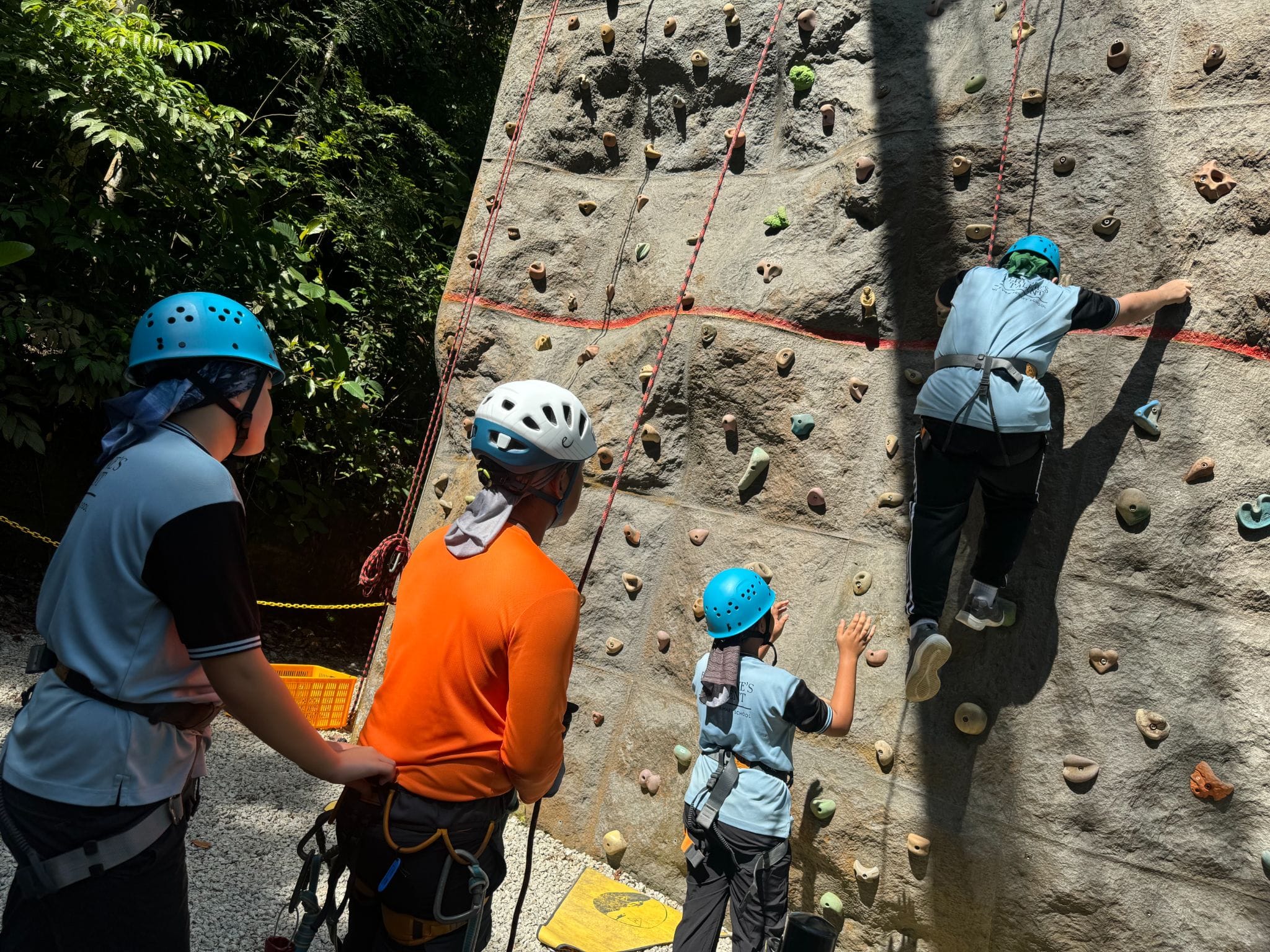 Four children wearing helmets and harnesses at an outdoor rock climbing wall, one climbing while others watch.