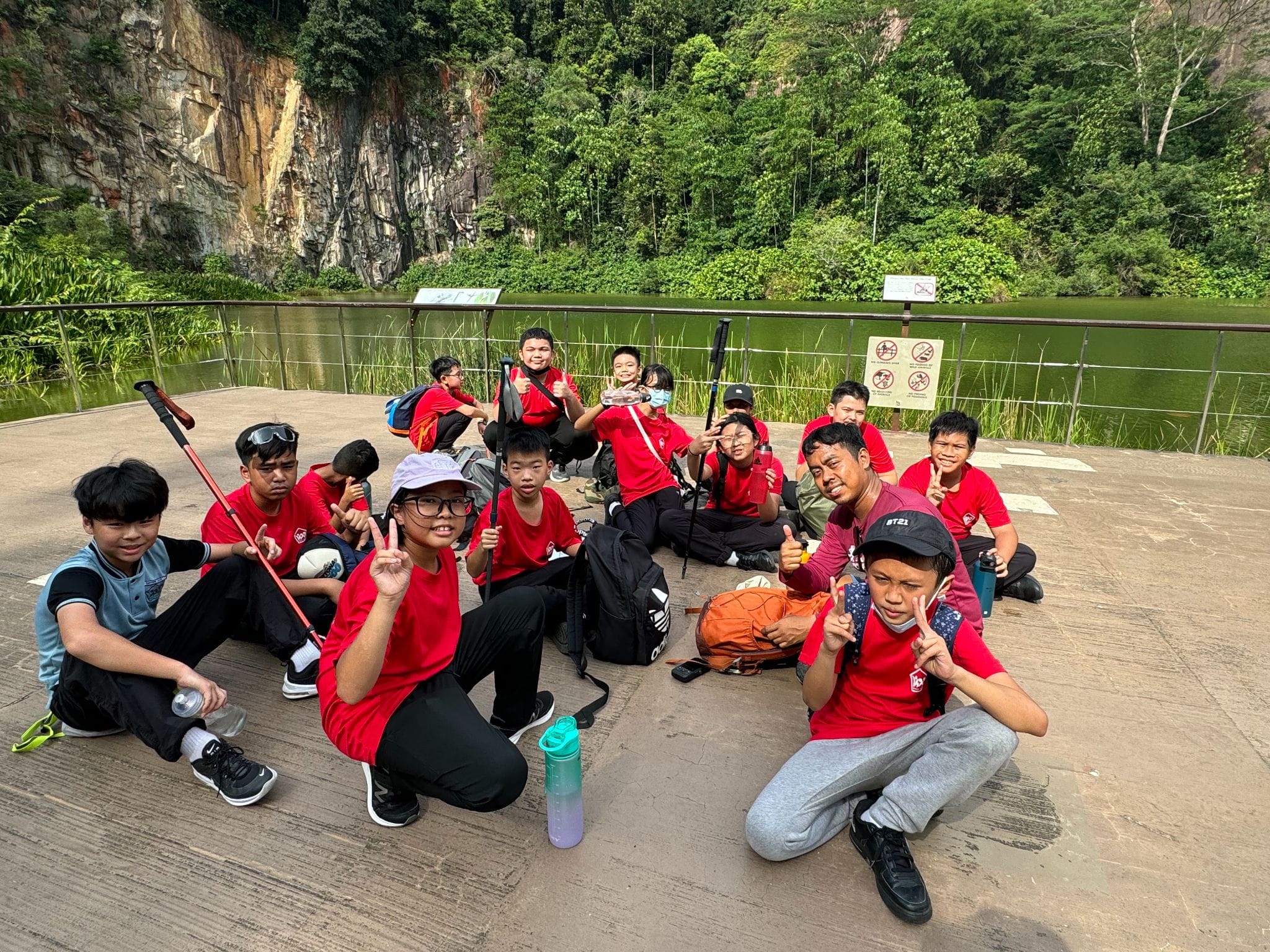 A group of children in red shirts pose and smile on a platform near a green lake with rocky cliffs behind them.