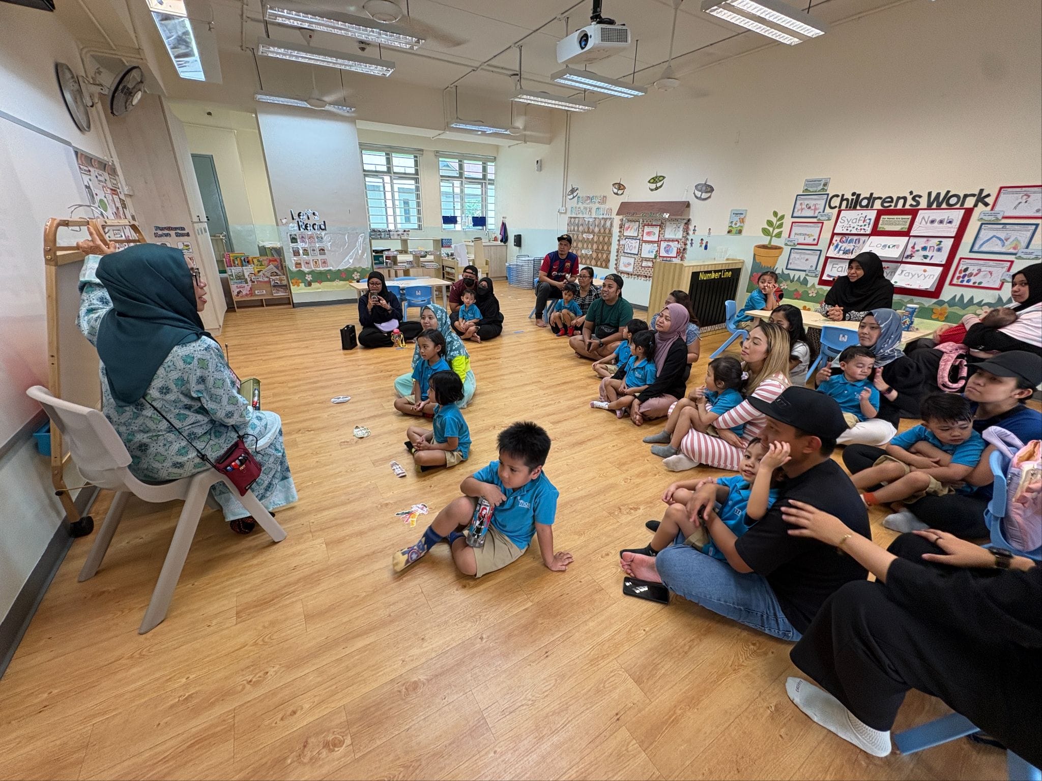 A teacher in a hijab sits facing young children and parents seated on a classroom floor near a Children's Work display.