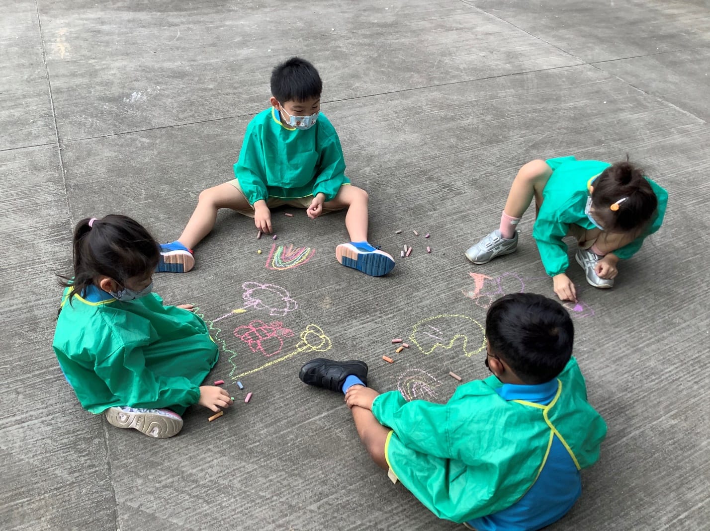 Four young children in green smocks and masks sit on concrete, drawing colorful chalk art together.