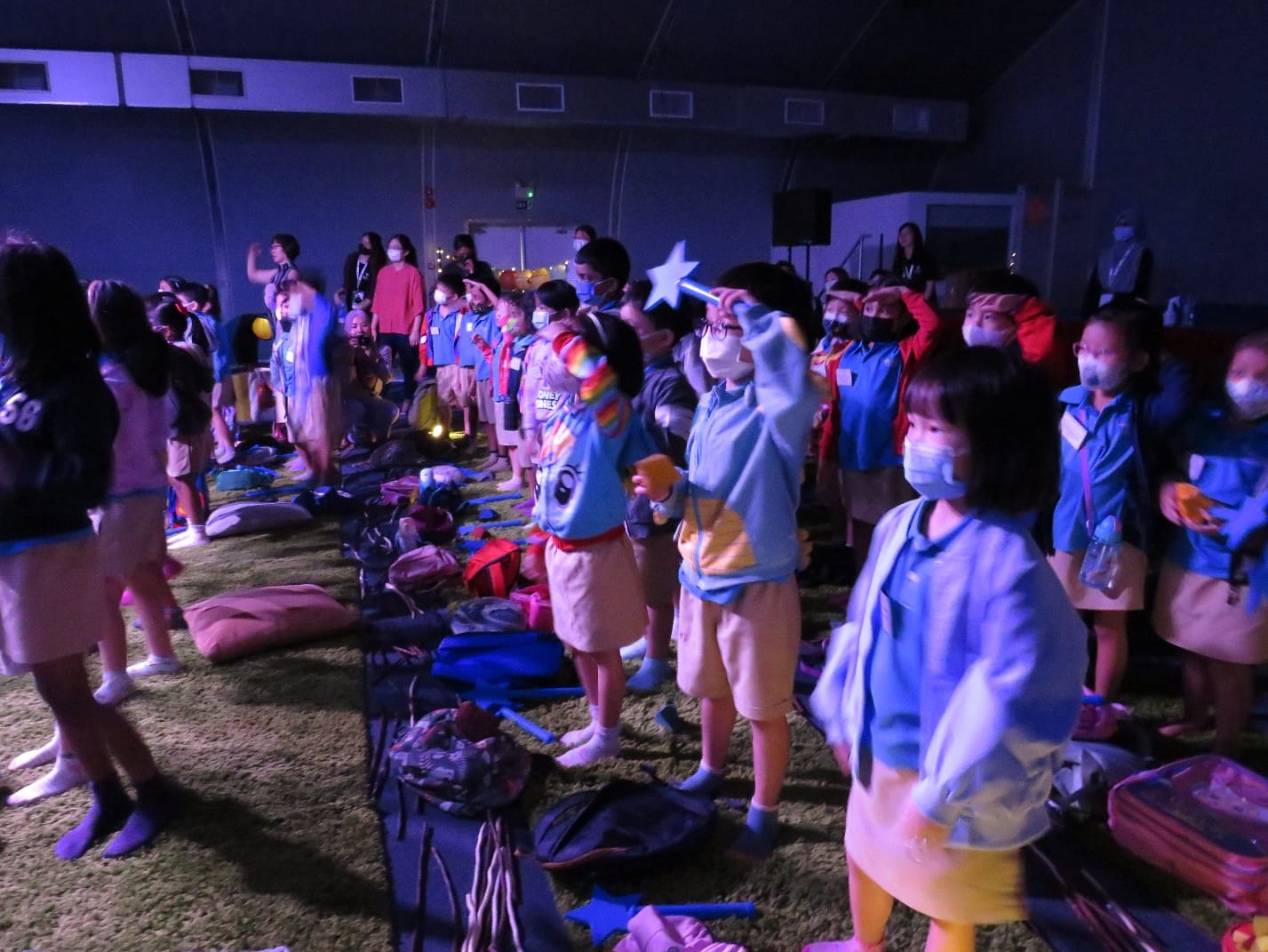 Children wearing masks and blue uniforms stand in a dimly lit indoor hall with bags on the floor.