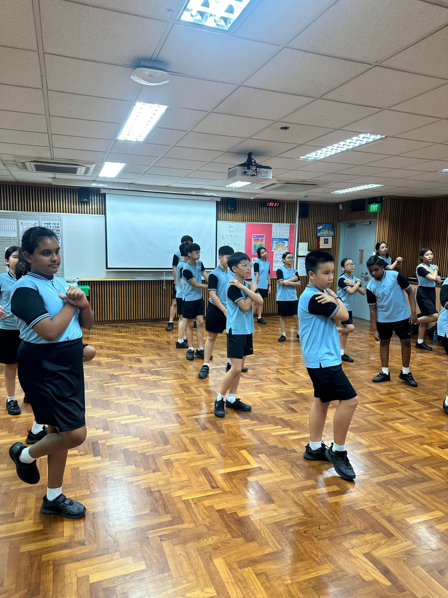 Students in blue and black uniforms practice a dance or exercise routine on a wooden floor in a school hall.