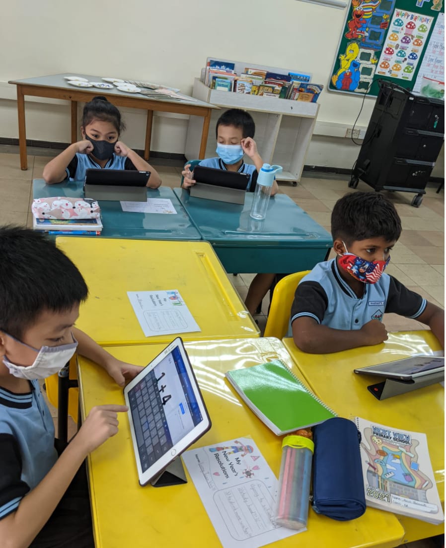 Four masked primary school children in blue uniforms use tablets at colorful desks in a classroom.