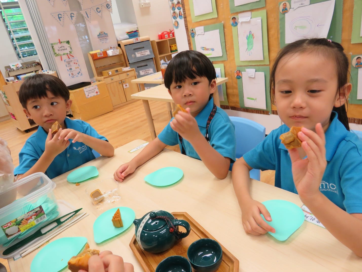 Three young children in blue school uniforms sit at a classroom table eating mooncakes with a teapot nearby.