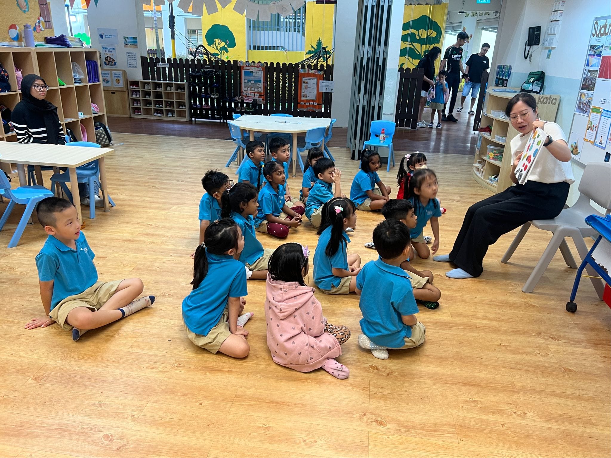 A teacher reads a book to young children in blue uniforms sitting on a classroom floor.