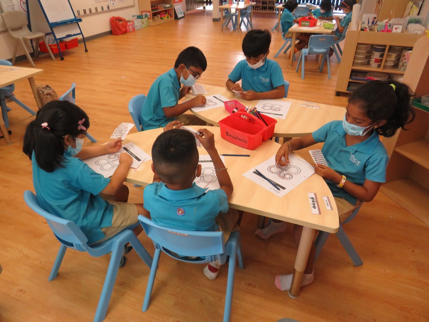 Five young children in teal uniforms and face masks sit around a table doing worksheet activities in a classroom.