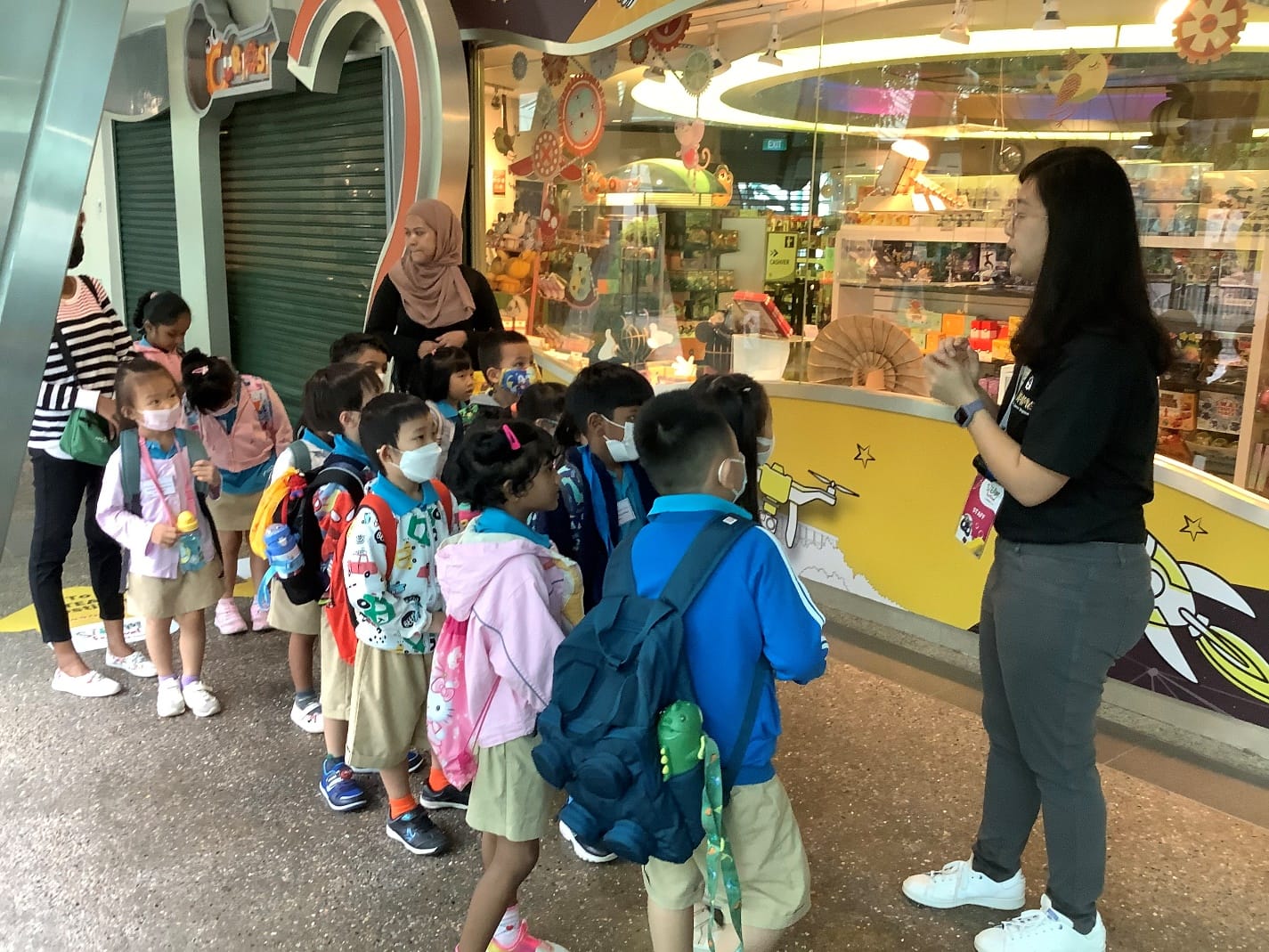 A female guide speaks to a group of young masked schoolchildren with backpacks outside a colorful toy shop.