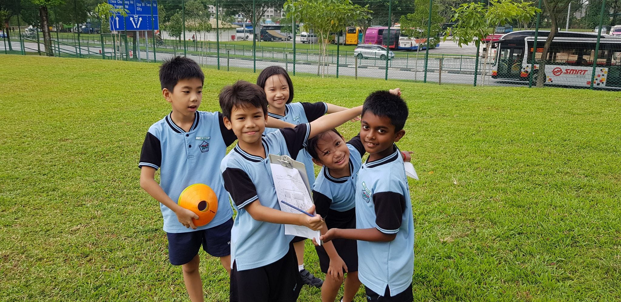 Five children in matching light blue school uniforms stand together smiling on a grass field, one holding an orange ball.